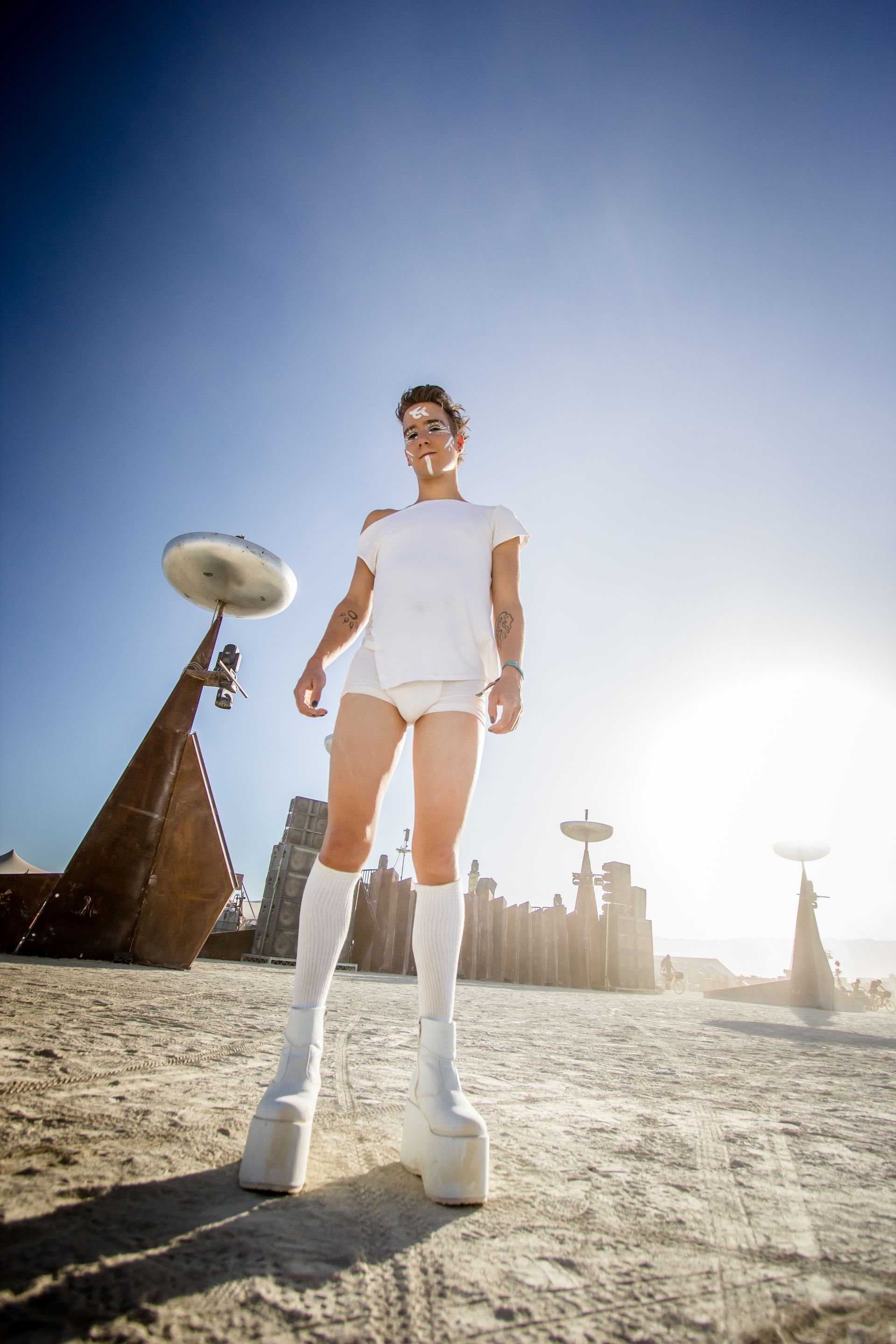 Person in white outfit and boots standing in a desert-like landscape with bright sun and futuristic structures