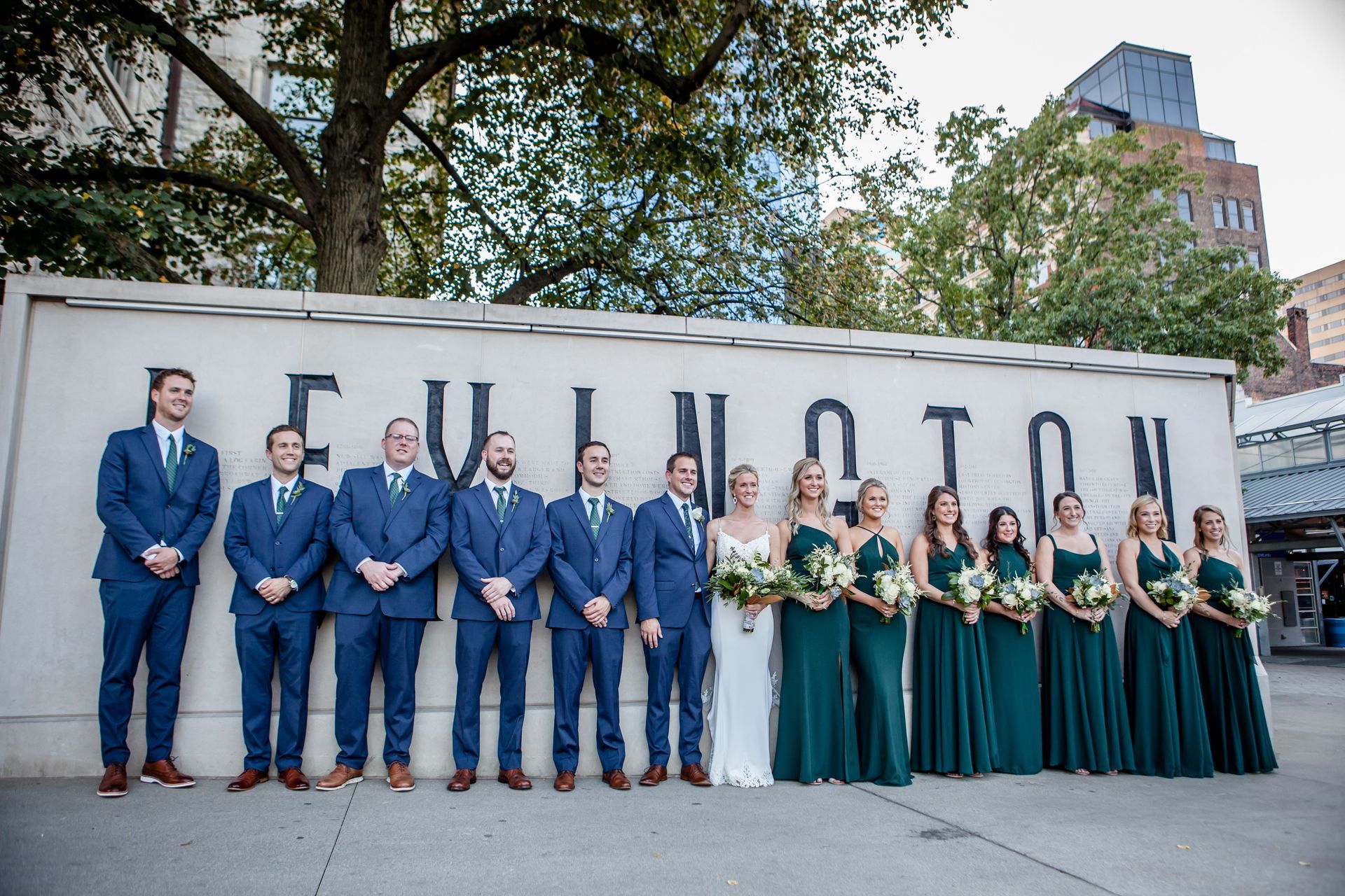 Wedding party in blue suits and green dresses standing in front of a FULTON sign outdoors