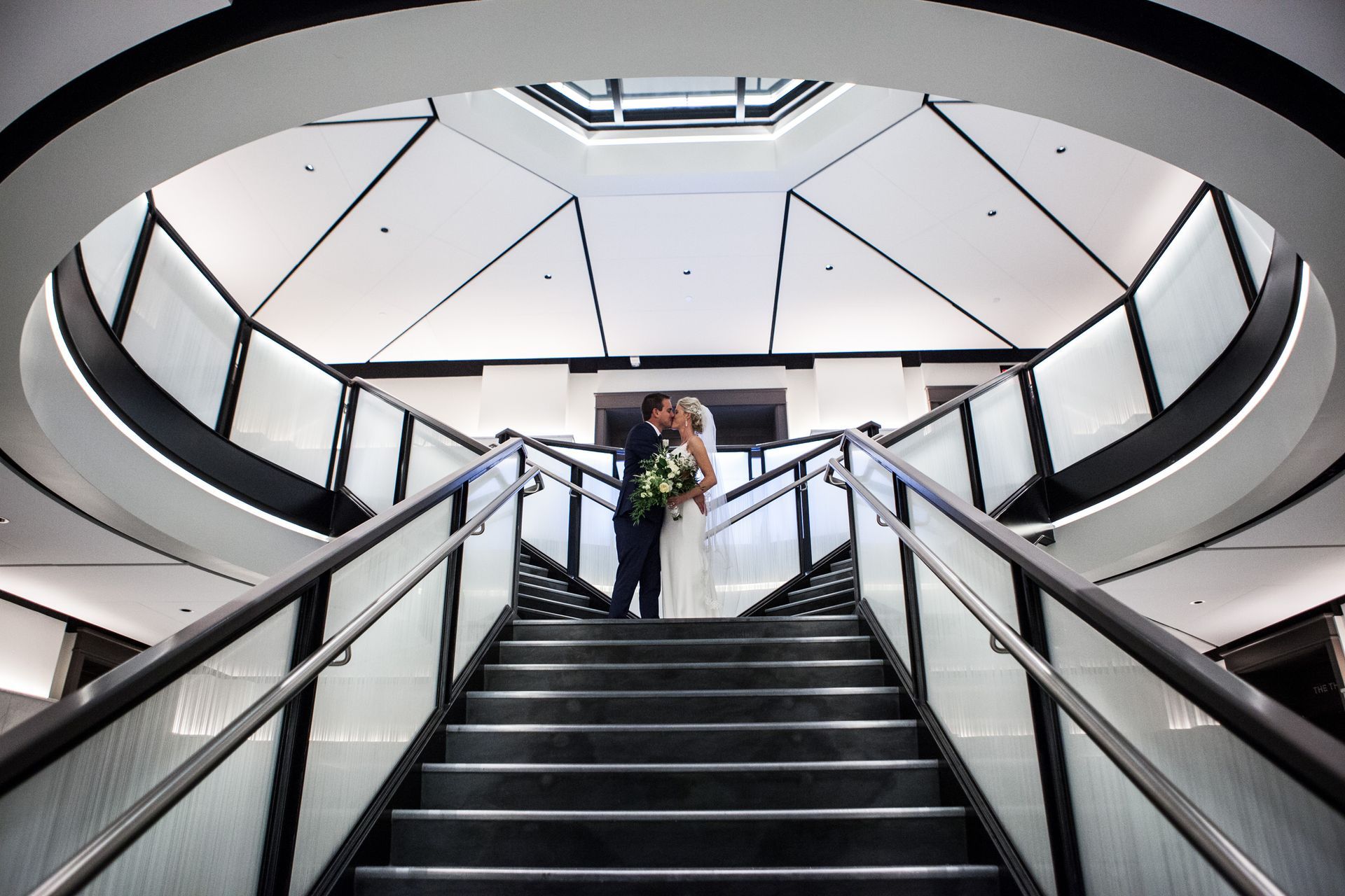 Wide staircase in a modern atrium with two people standing at the top under a geometric ceiling.
