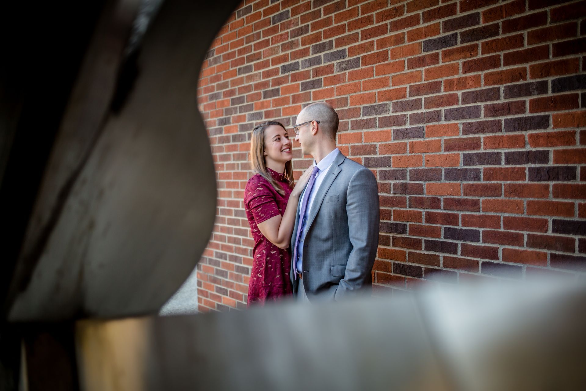 Couple standing close together by a brick wall, with a blurred foreground and soft natural light.