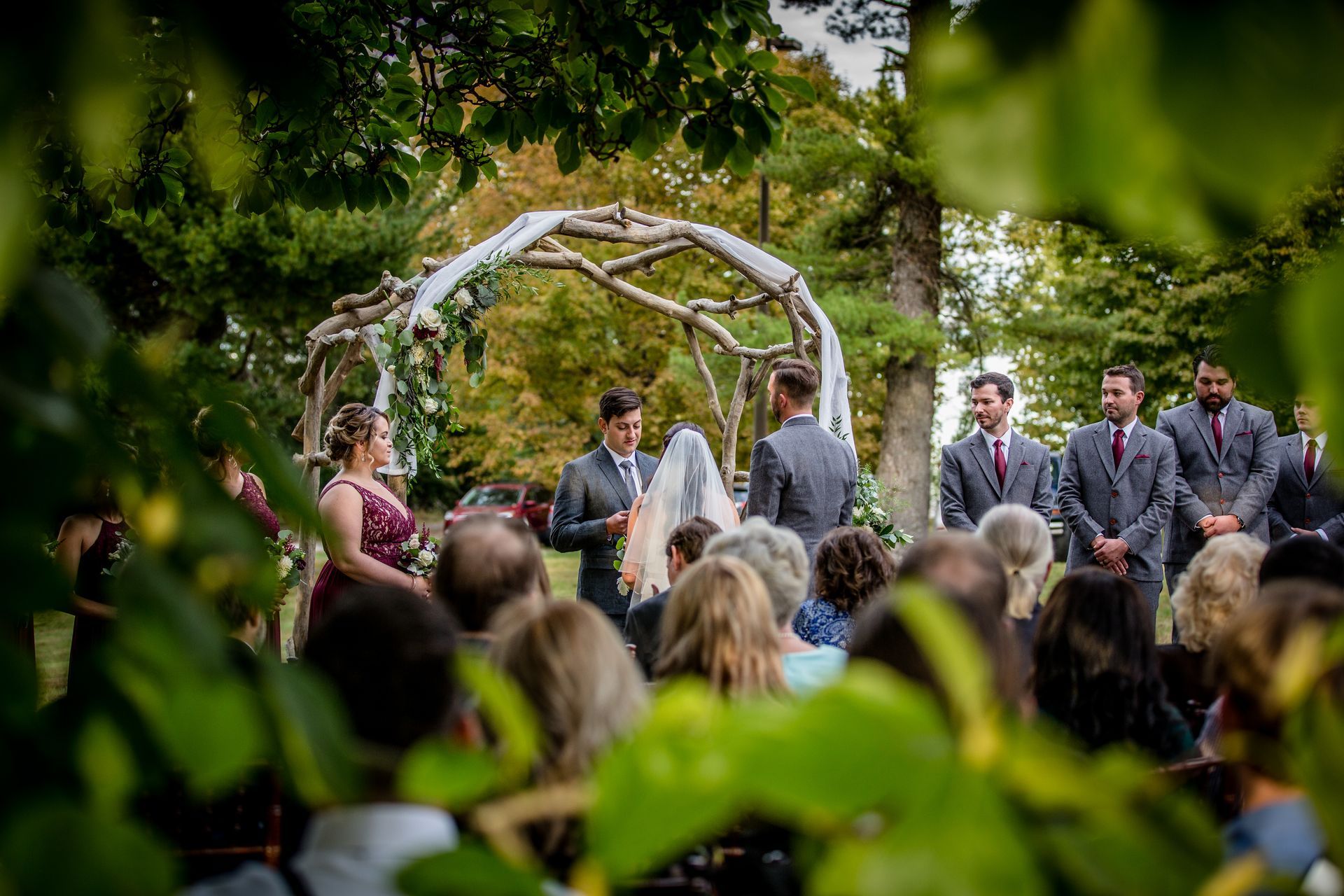 Outdoor wedding ceremony with wedding party under a floral arch, guests seated in the foreground