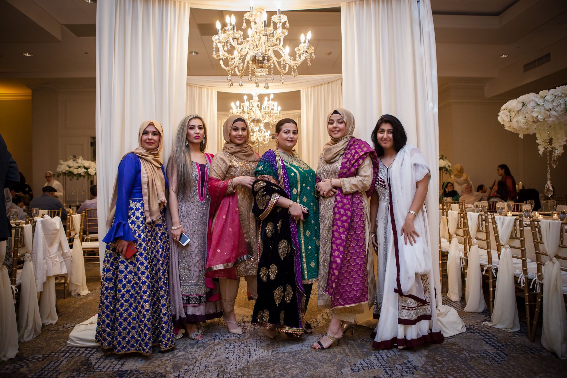 Six women in colorful dresses pose together in a decorated banquet hall under a chandelier.