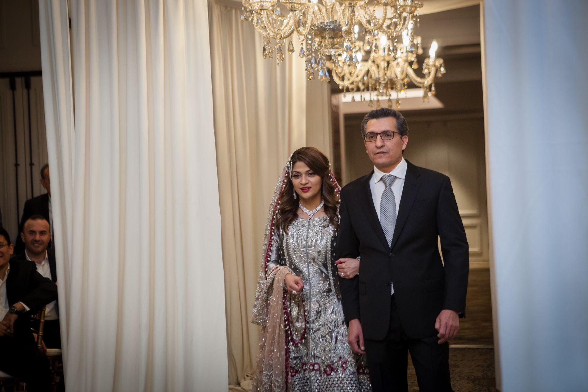 Two people in formal attire walking through a chandelier-lit hall with curtains