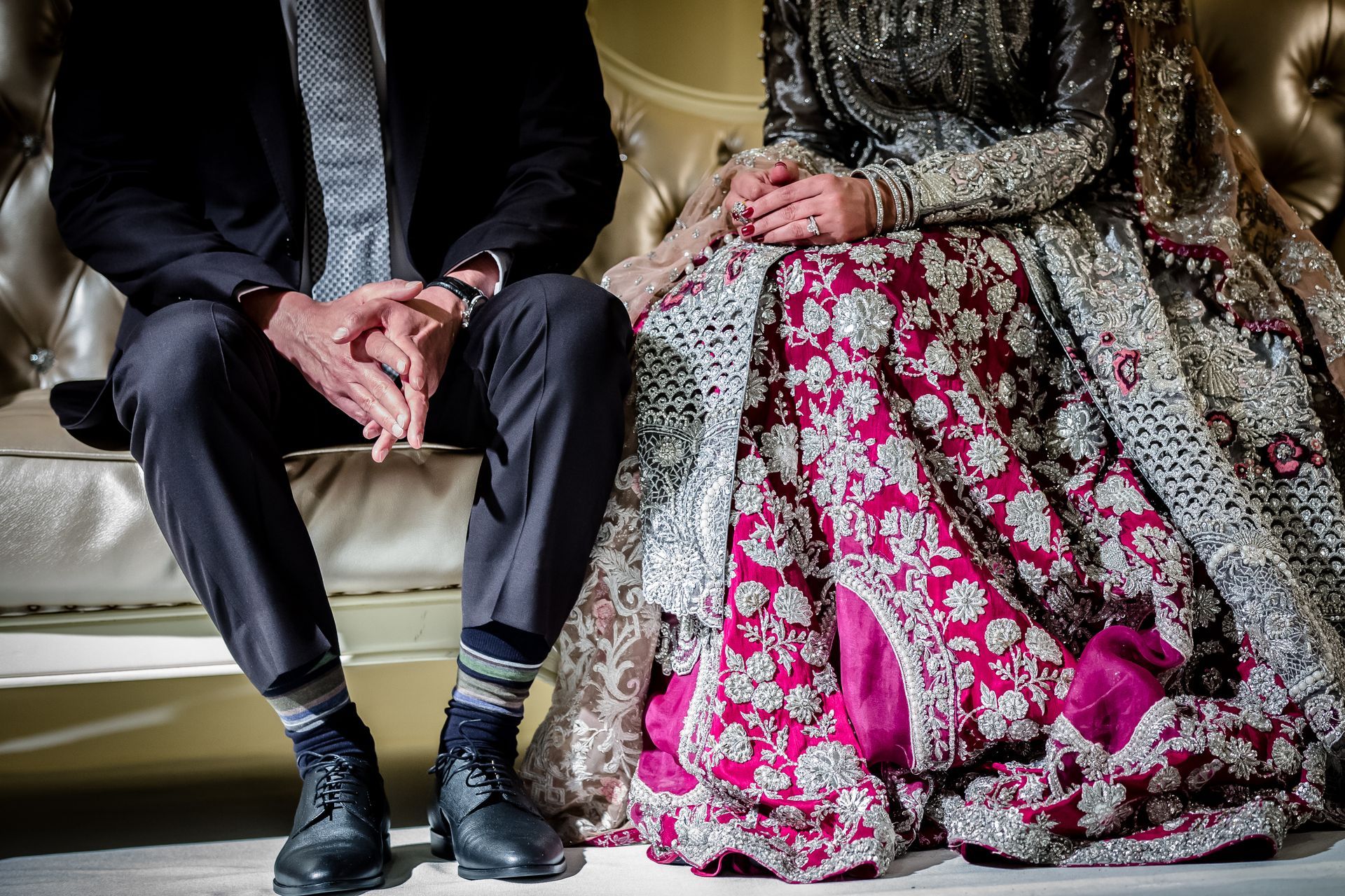 Two seated people in formal attire on a cream sofa, hands folded, with a bright pink floral skirt in the center