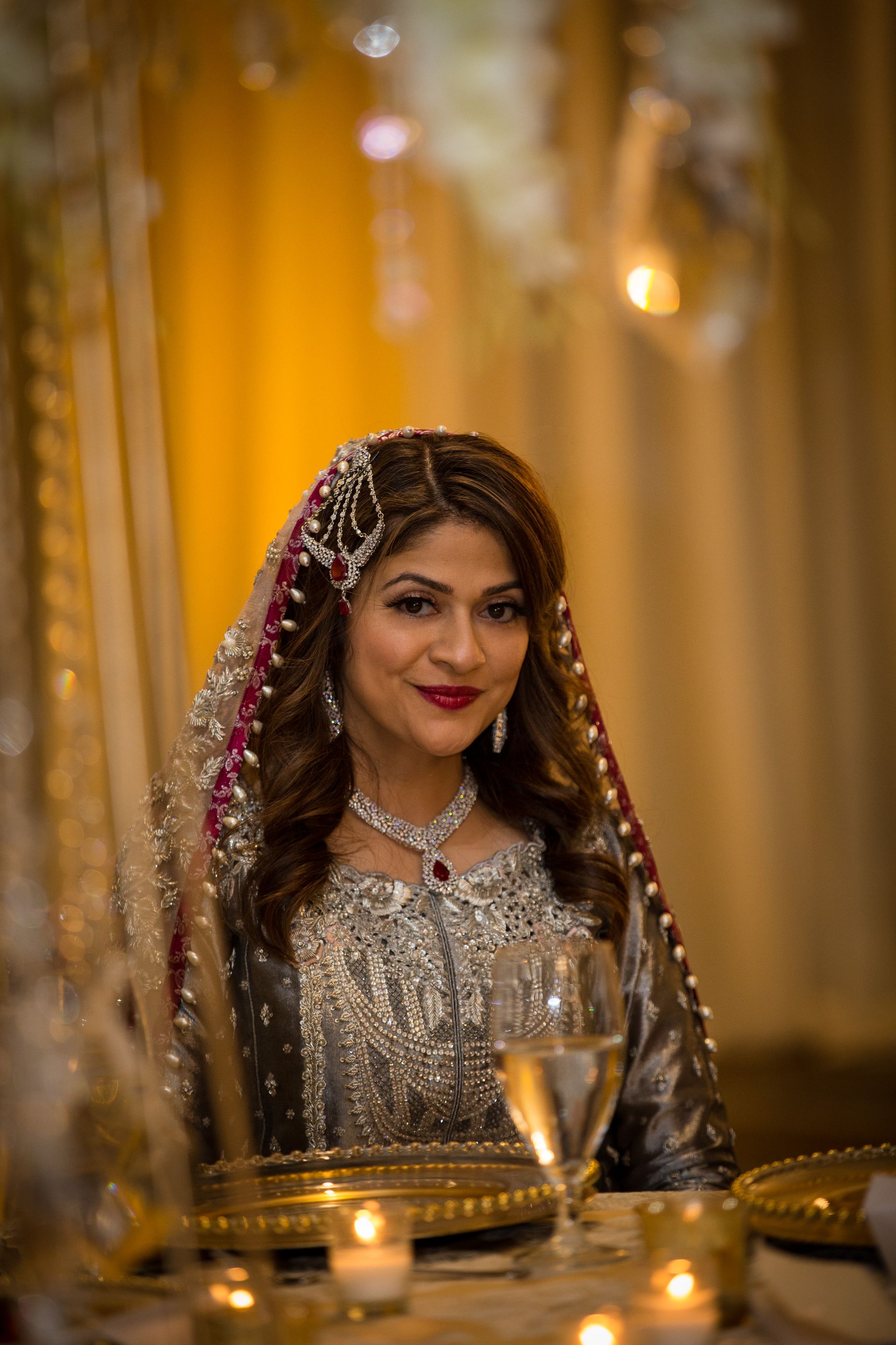 Woman in silver traditional dress and pink veil seated at a candlelit table, smiling at the camera.