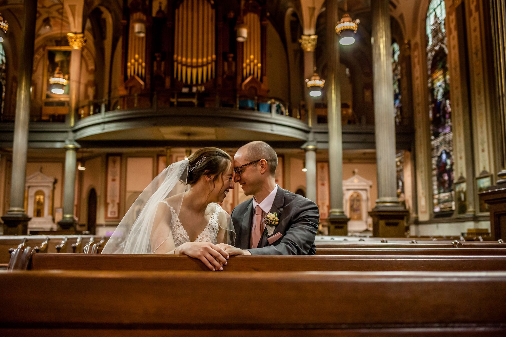 Bride and groom seated in a cathedral pew, leaning close beneath ornate arches.