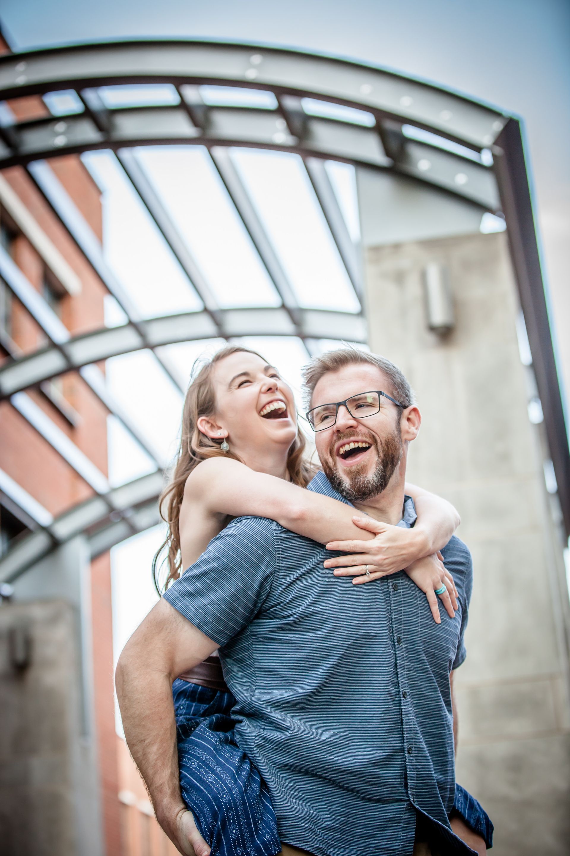 Smiling couple outdoors, laughing and embracing beneath a curved glass canopy