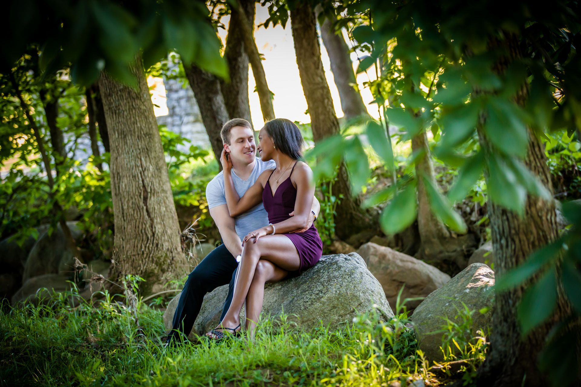Two people sitting on a rock in a sunlit forest, leaning close together.