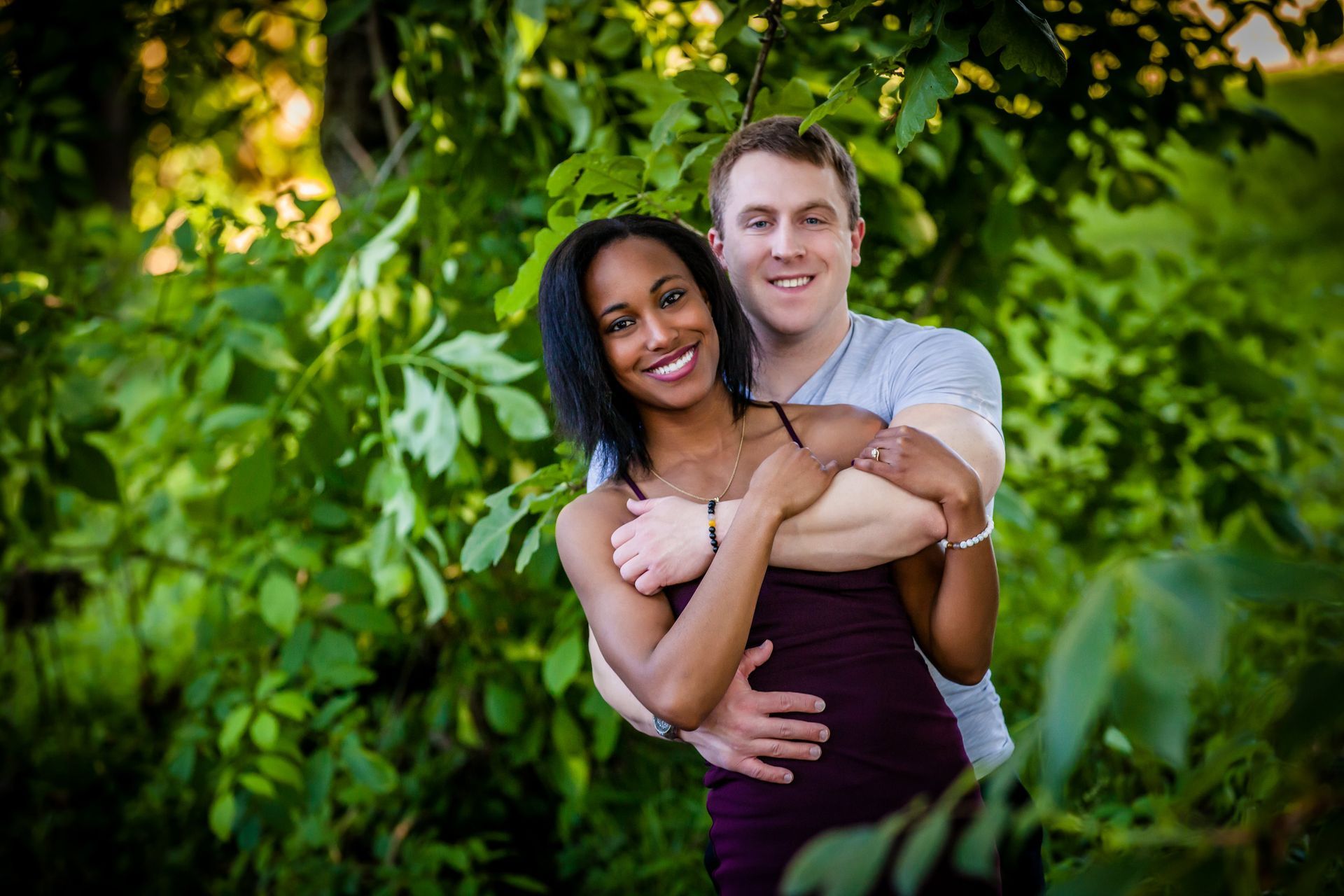 Smiling couple embracing in a leafy garden, with green foliage and soft sunlight.