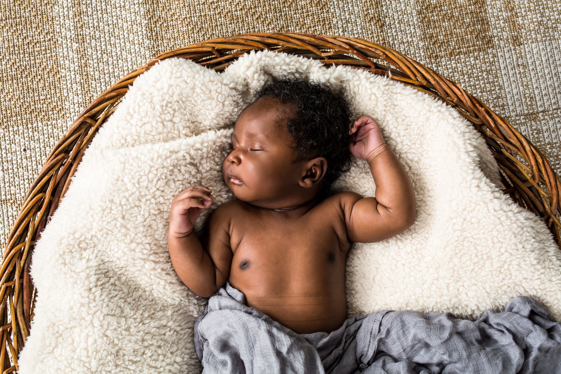 Sleeping baby on a cream blanket in a woven basket, wearing gray pants and resting with one hand near the head