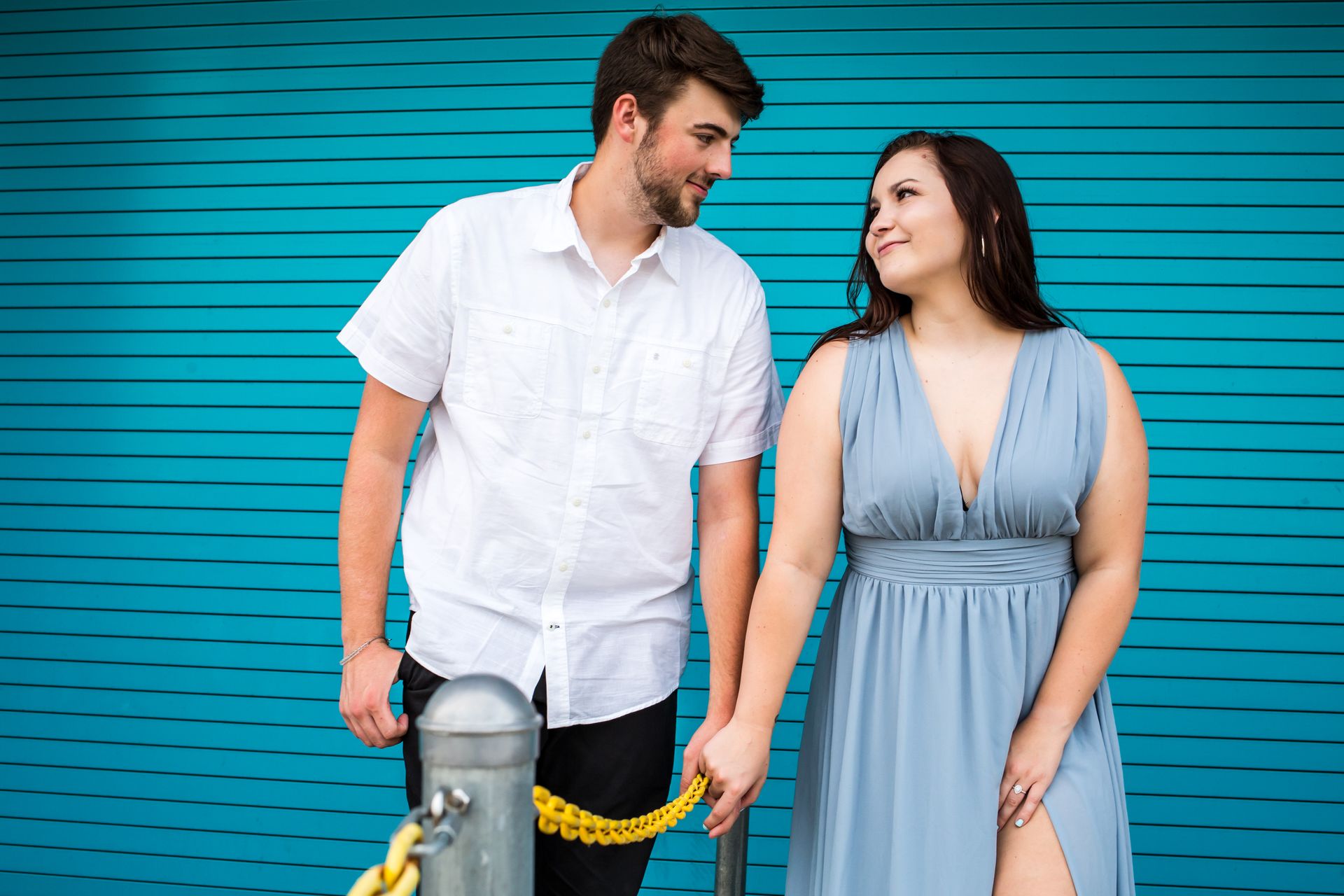 Two people stand smiling beside a yellow rope barrier in front of a turquoise wall.