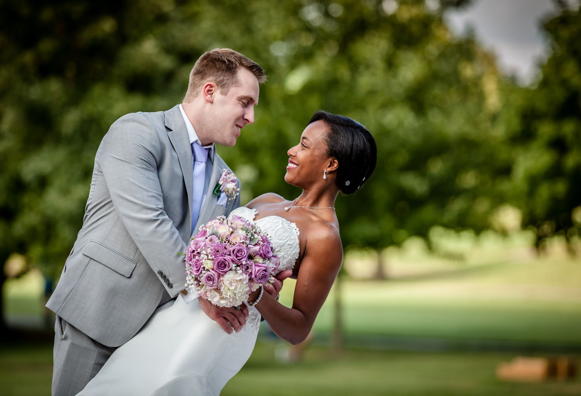 Bride and groom in wedding attire smiling and leaning together outdoors, holding a pink bouquet