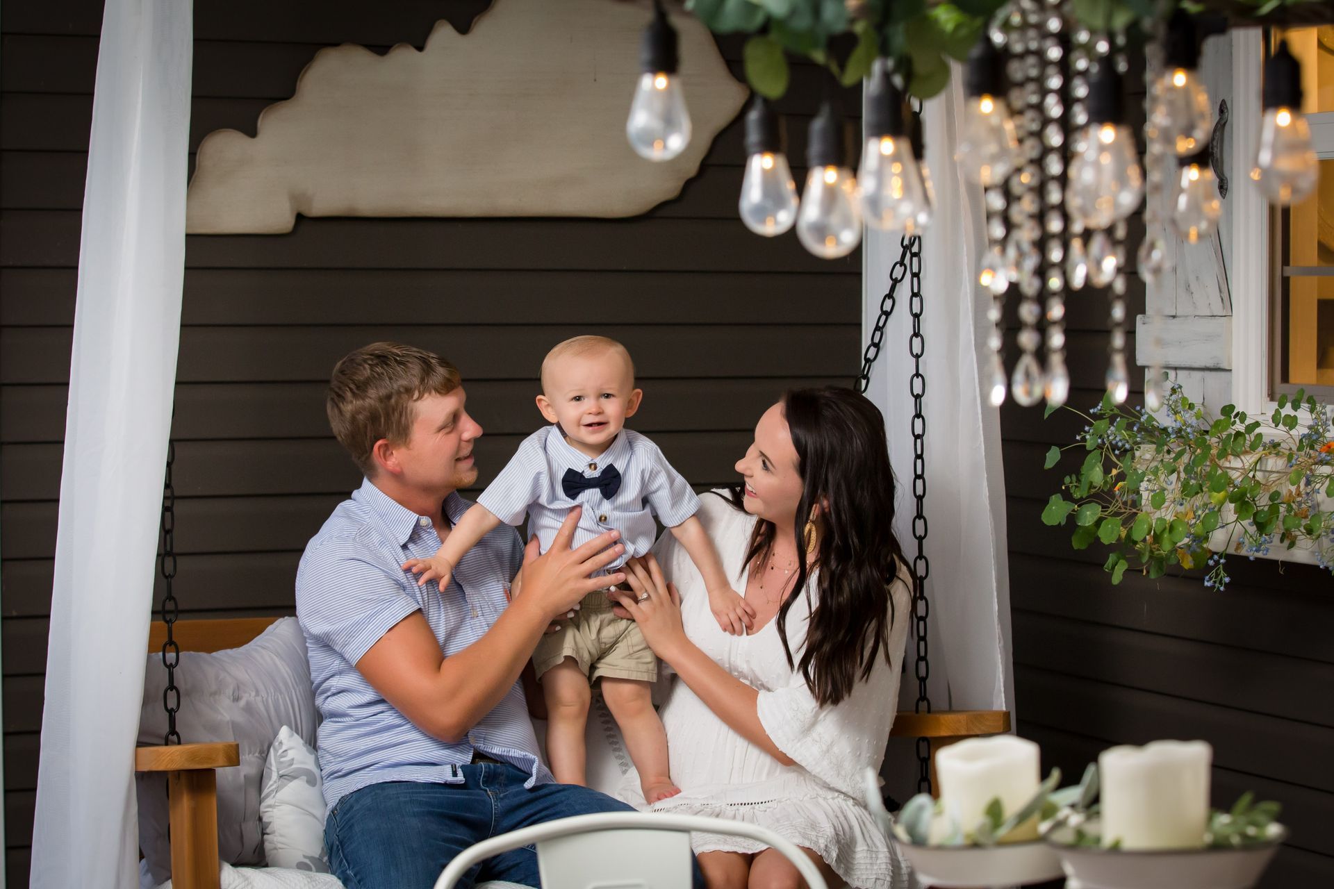 Family sitting on a porch swing, smiling at a baby under hanging lights and flowers.