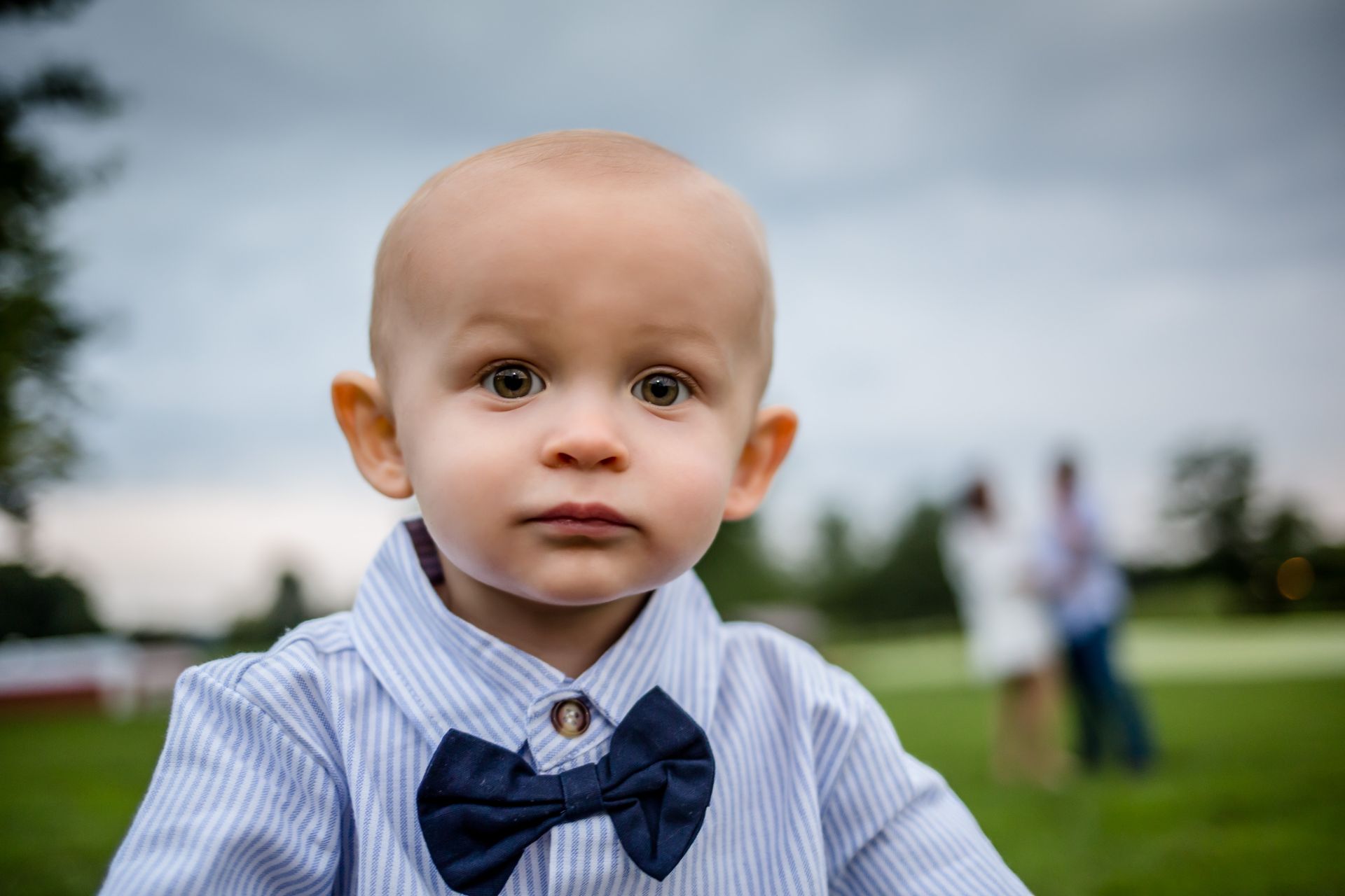 Child in a blue striped shirt and bow tie standing outdoors on a grassy field.
