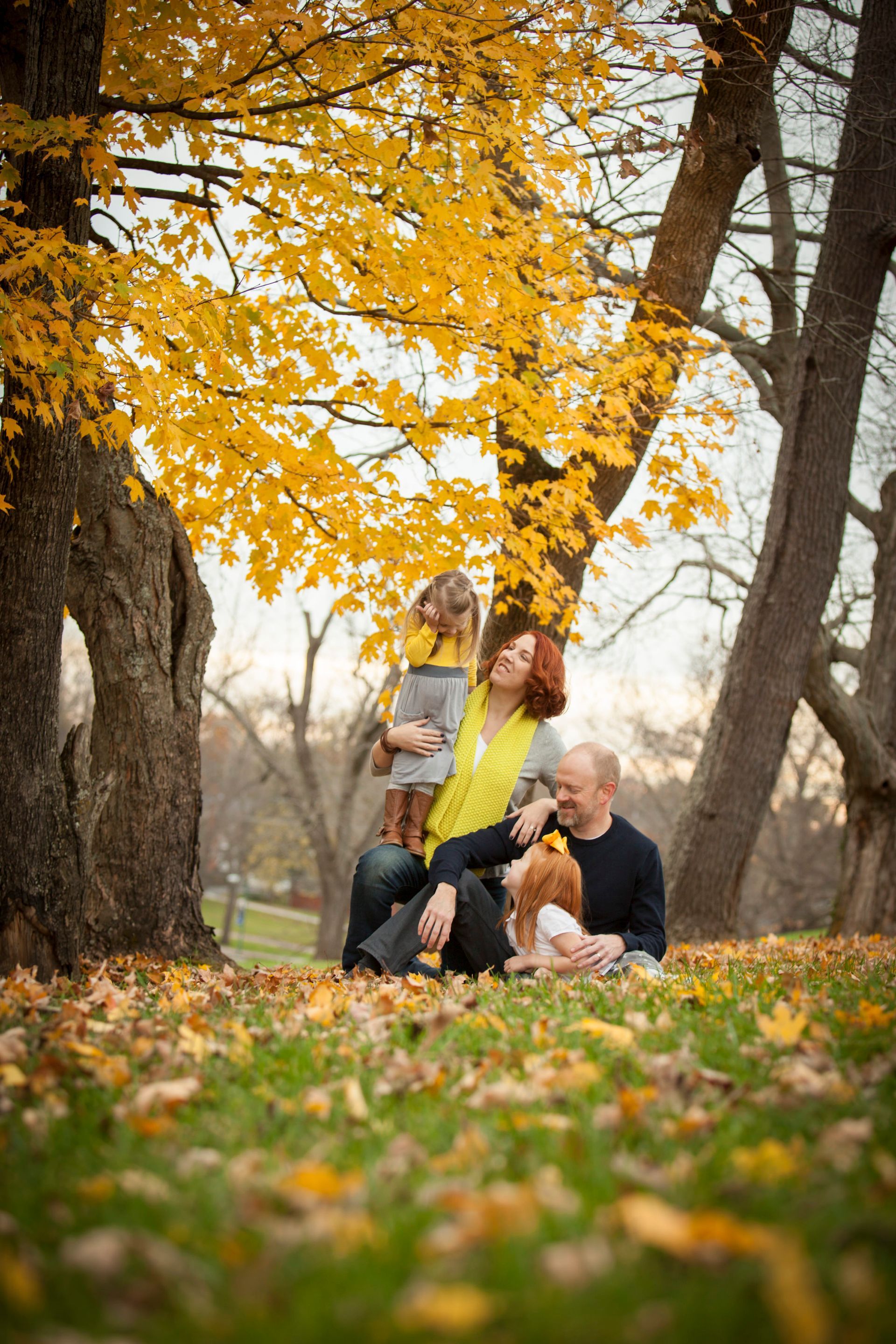 Family sitting on autumn leaves under yellow trees in a park, with a child and dog nearby