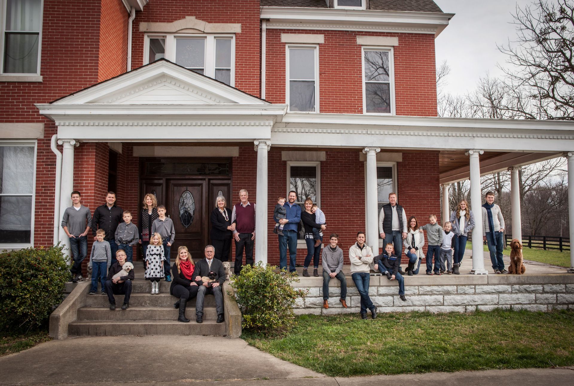 Large group posing on a porch and steps in front of a red brick house with white columns