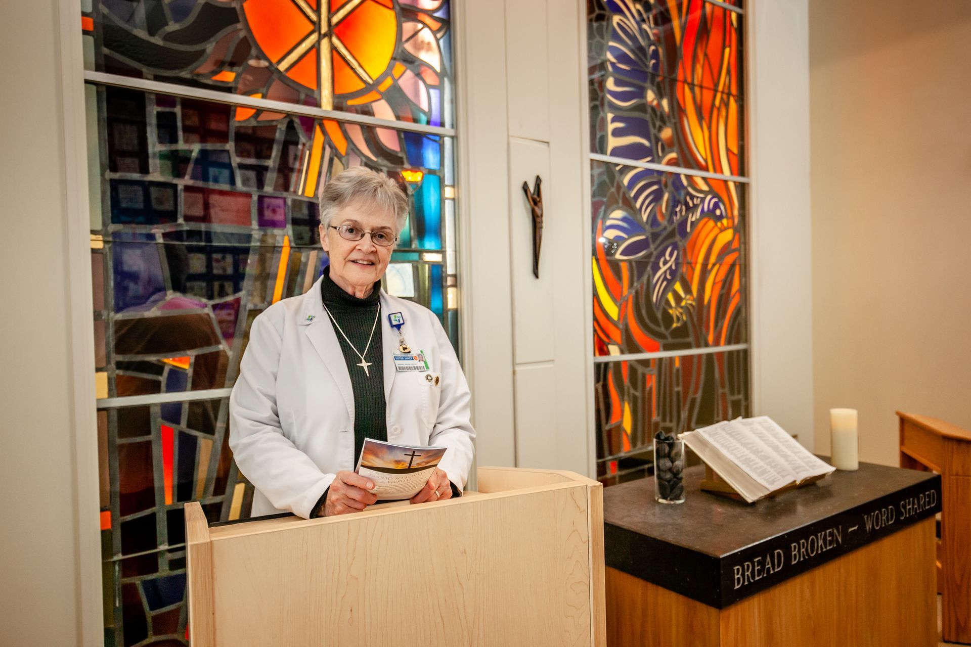 Person in a white coat stands at a lectern beside stained-glass windows in a chapel.