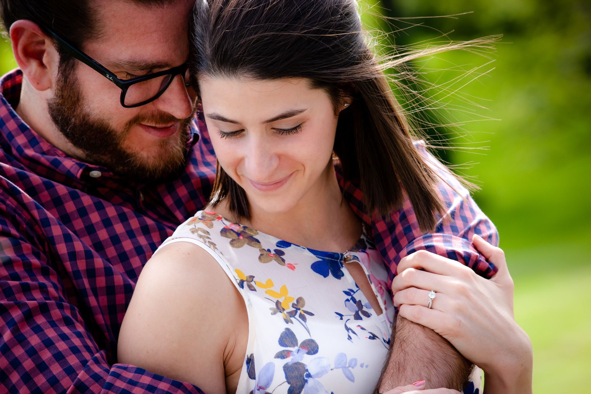 Couple embracing outdoors, the woman smiling in a white floral dress and the man in glasses and a plaid shirt.