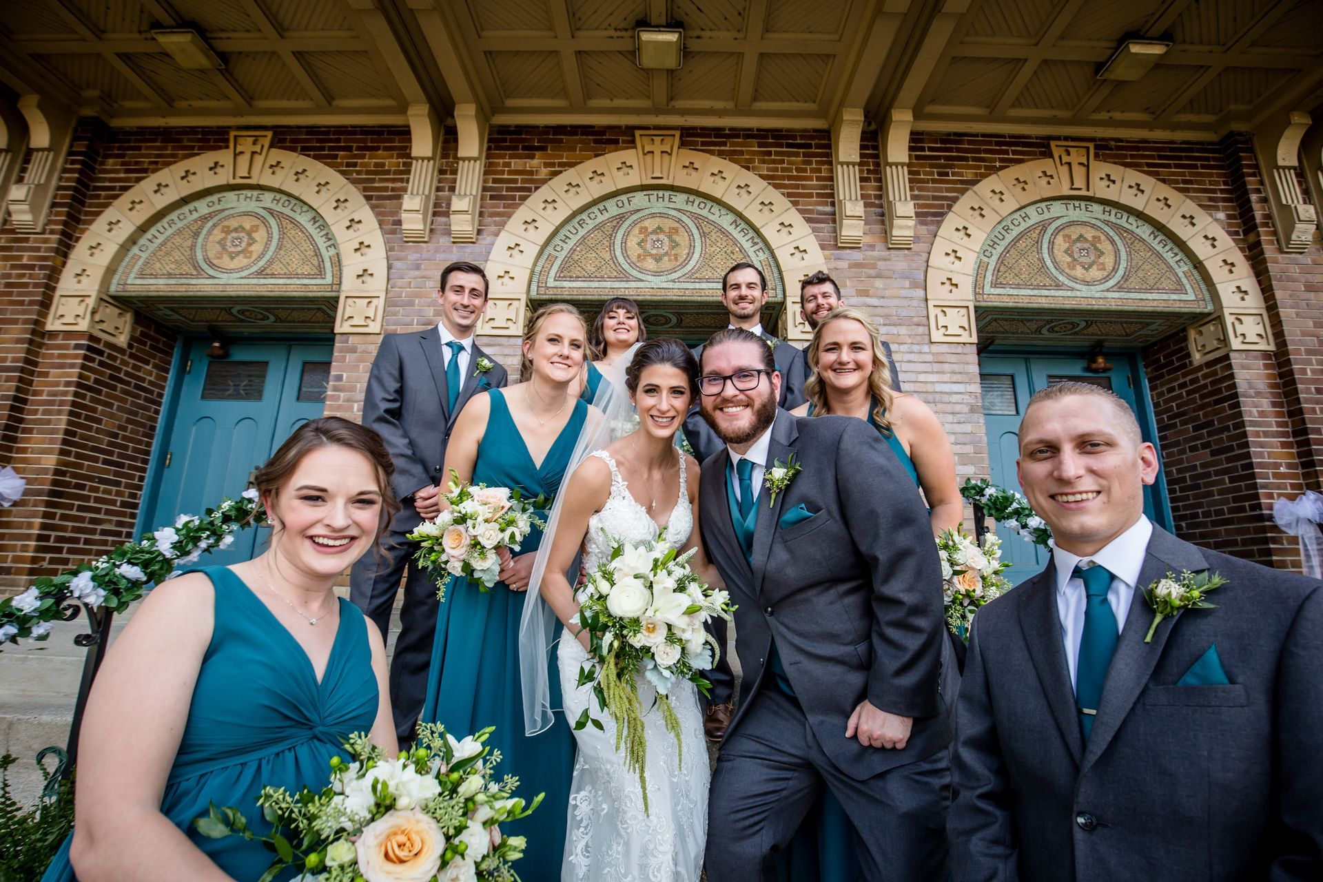 Wedding party posing in front of a brick building with arched doors, bouquets, and formal attire