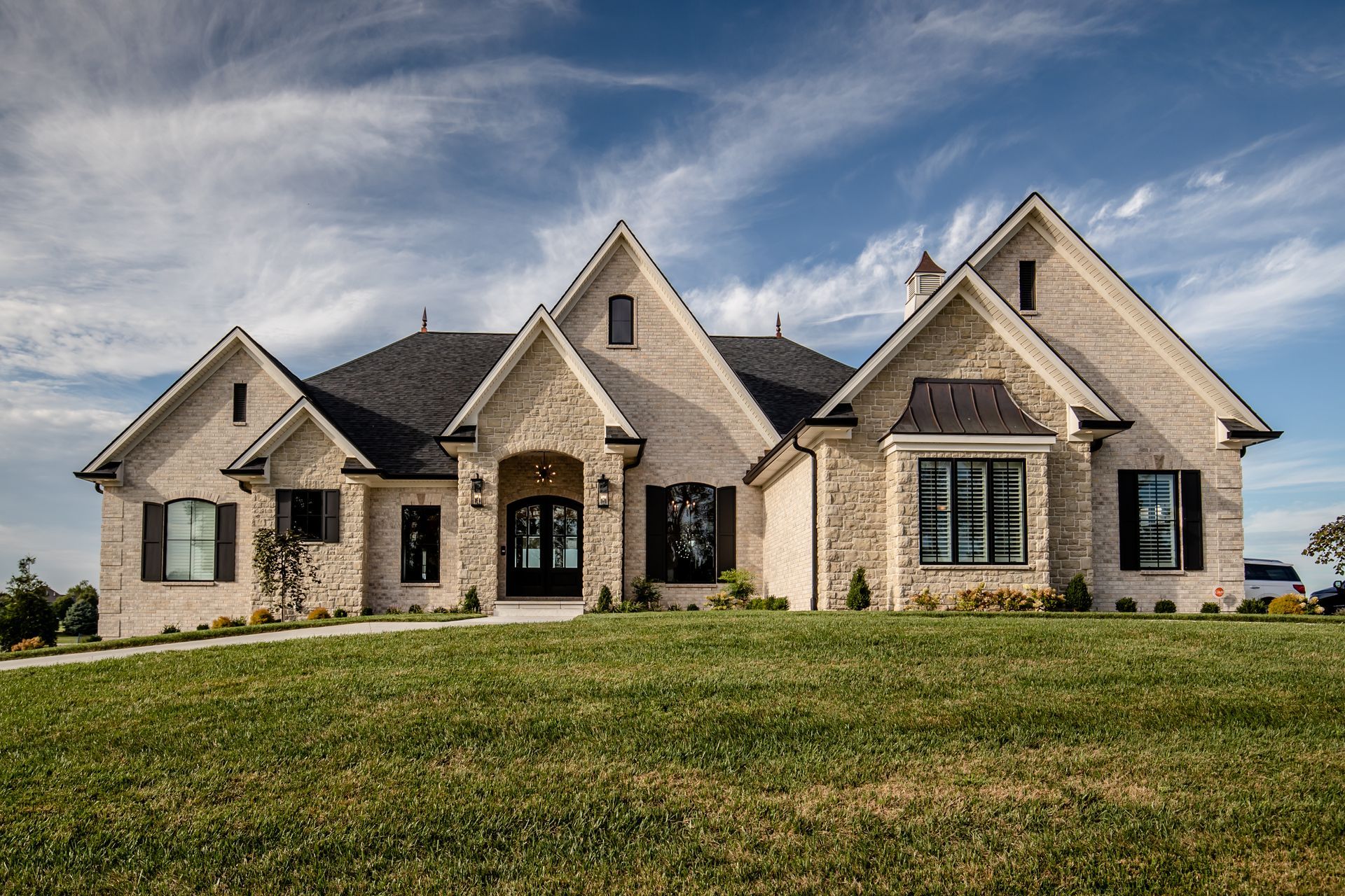 Large stone suburban house with multiple gables and a wide front lawn under a blue sky