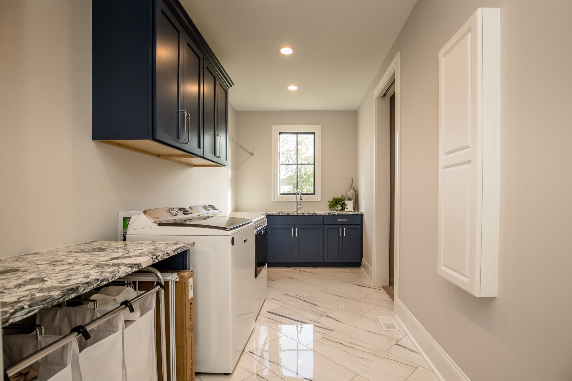 Narrow laundry room with navy cabinets, marble countertop, white washer, and glossy tile floor
