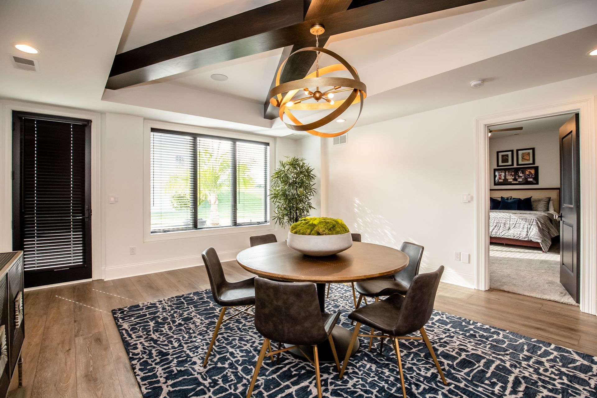 Modern dining room with round table, dark chairs, blue patterned rug, and exposed wood ceiling beams