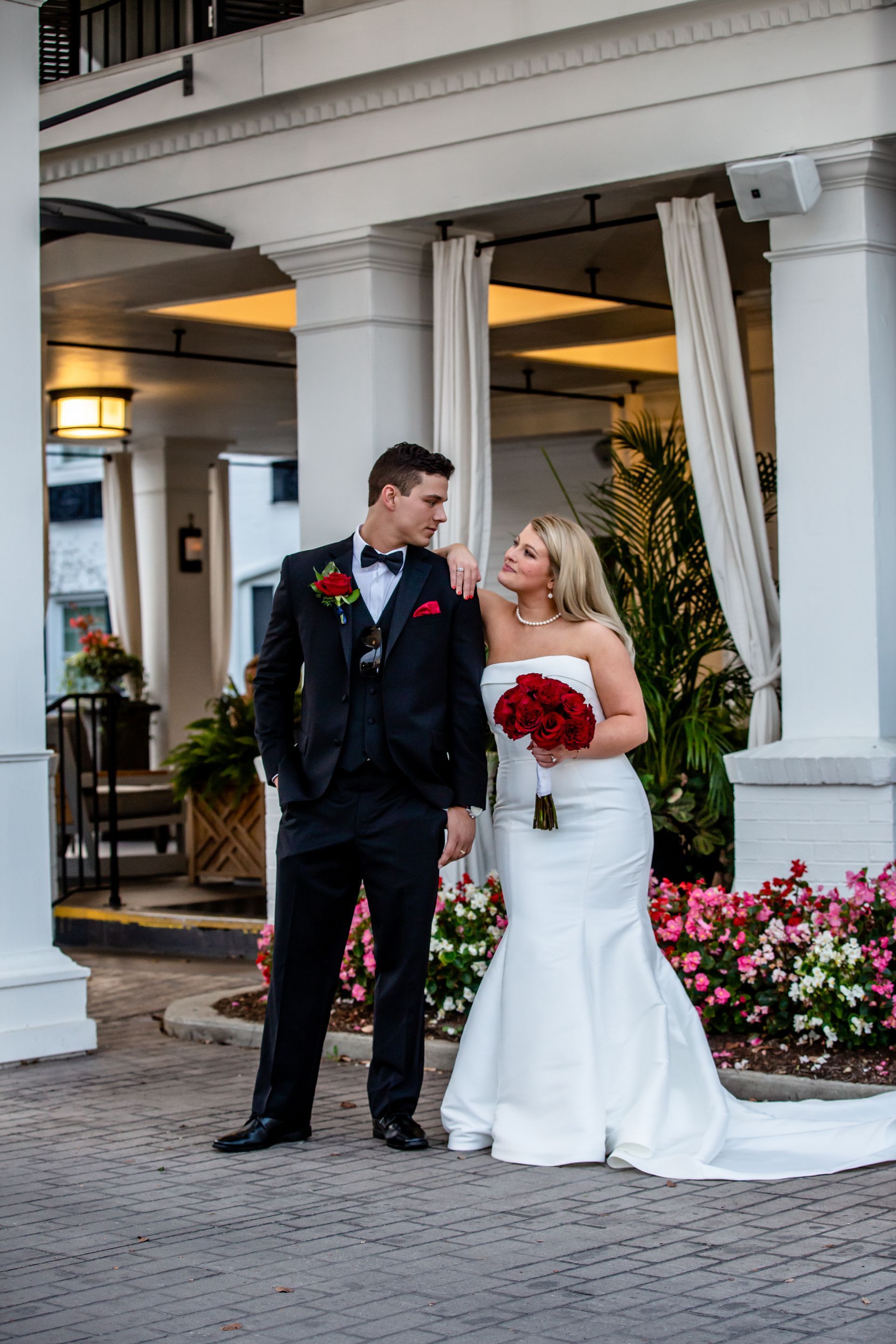 Bride and groom standing together outside a white-columned building, the bride holding a red bouquet.