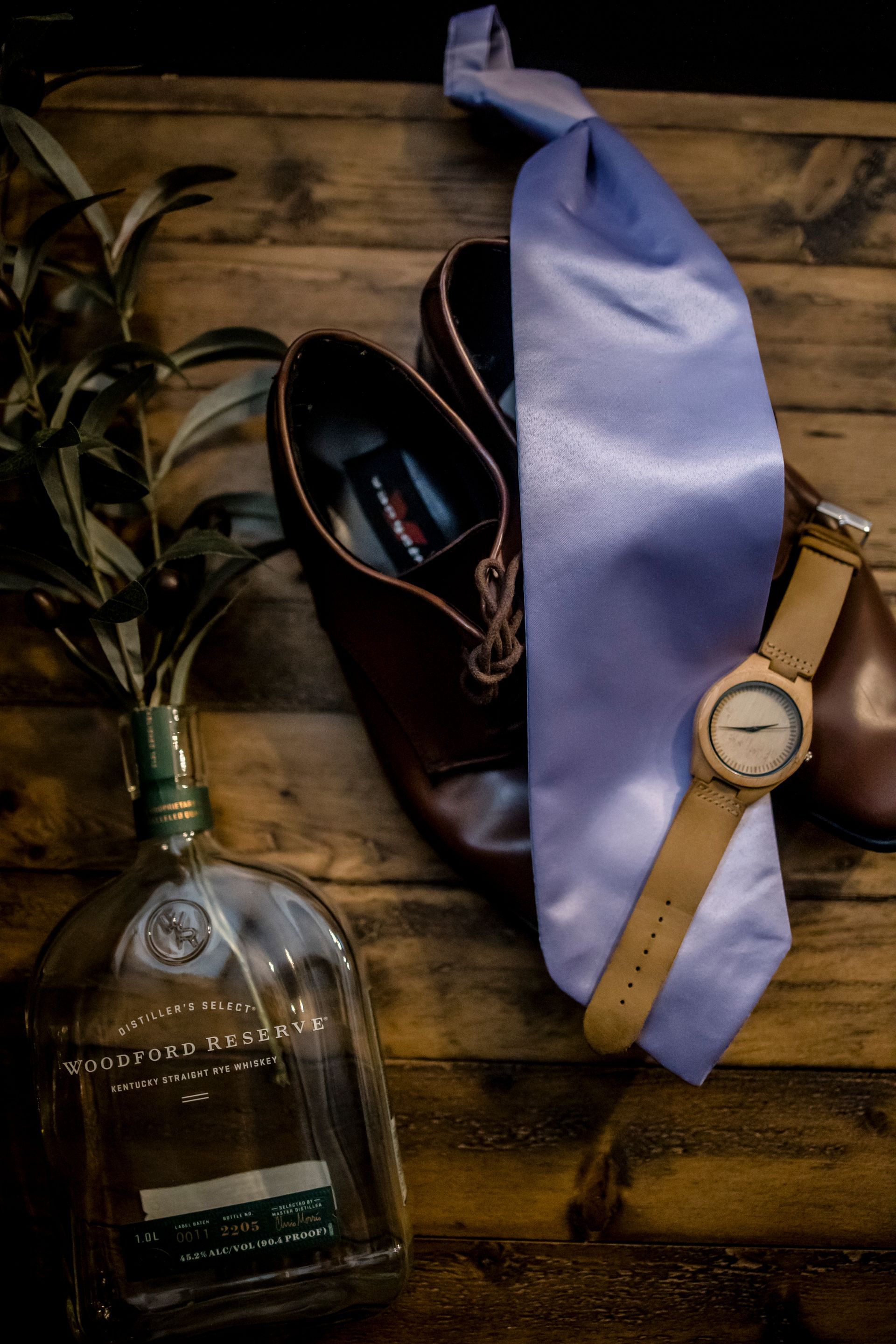 Tie, watch, shoes, and a liquor bottle arranged on a wooden surface.