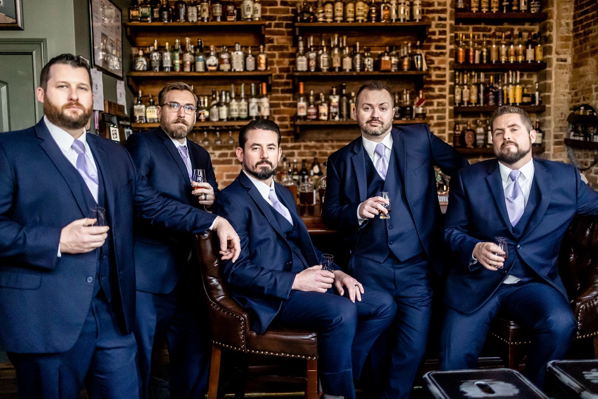 Five men in navy suits holding drinks in a bar with shelves of liquor behind them.