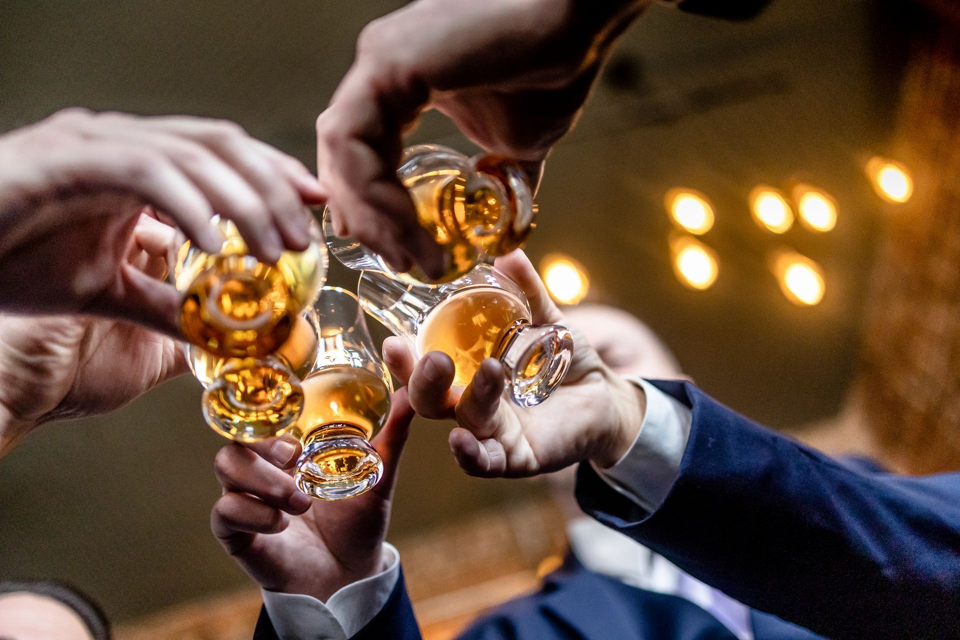Hands raising glasses in a toast, with amber drinks in a warm, blurred party setting