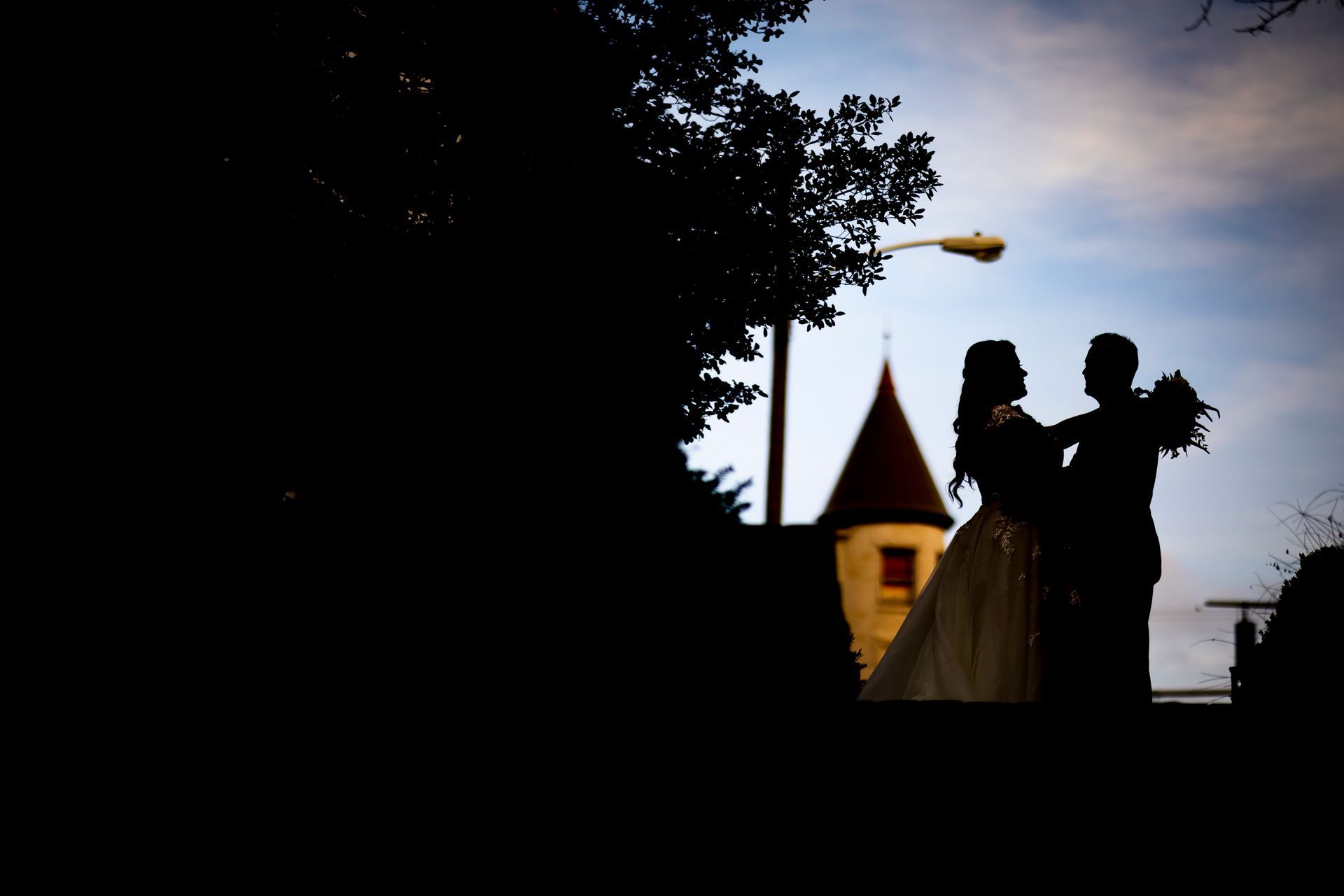 Silhouetted couple in wedding attire outdoors at dusk near a lit church tower