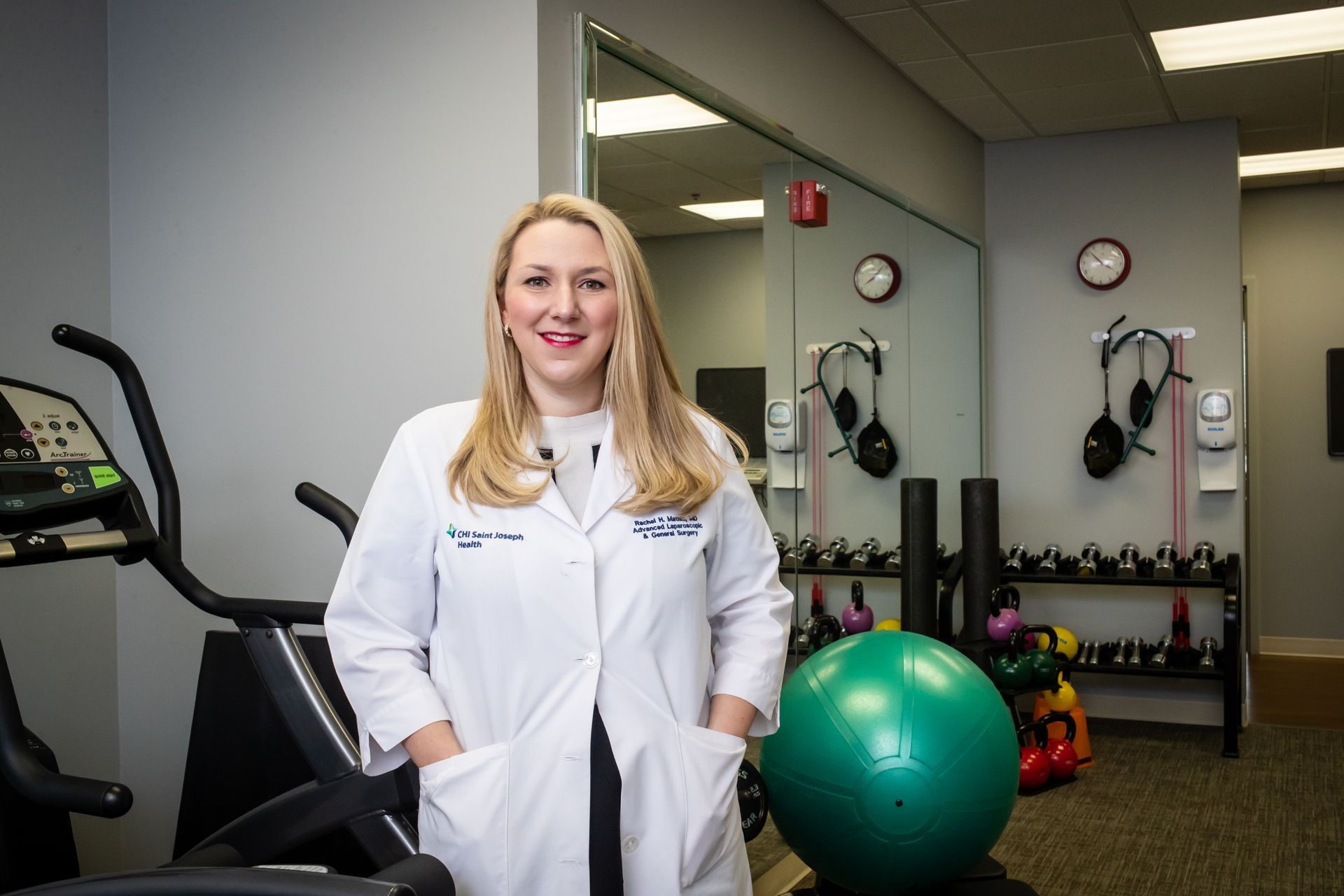 Healthcare worker in white lab coat standing in a gym or rehab room with exercise equipment and a stability ball