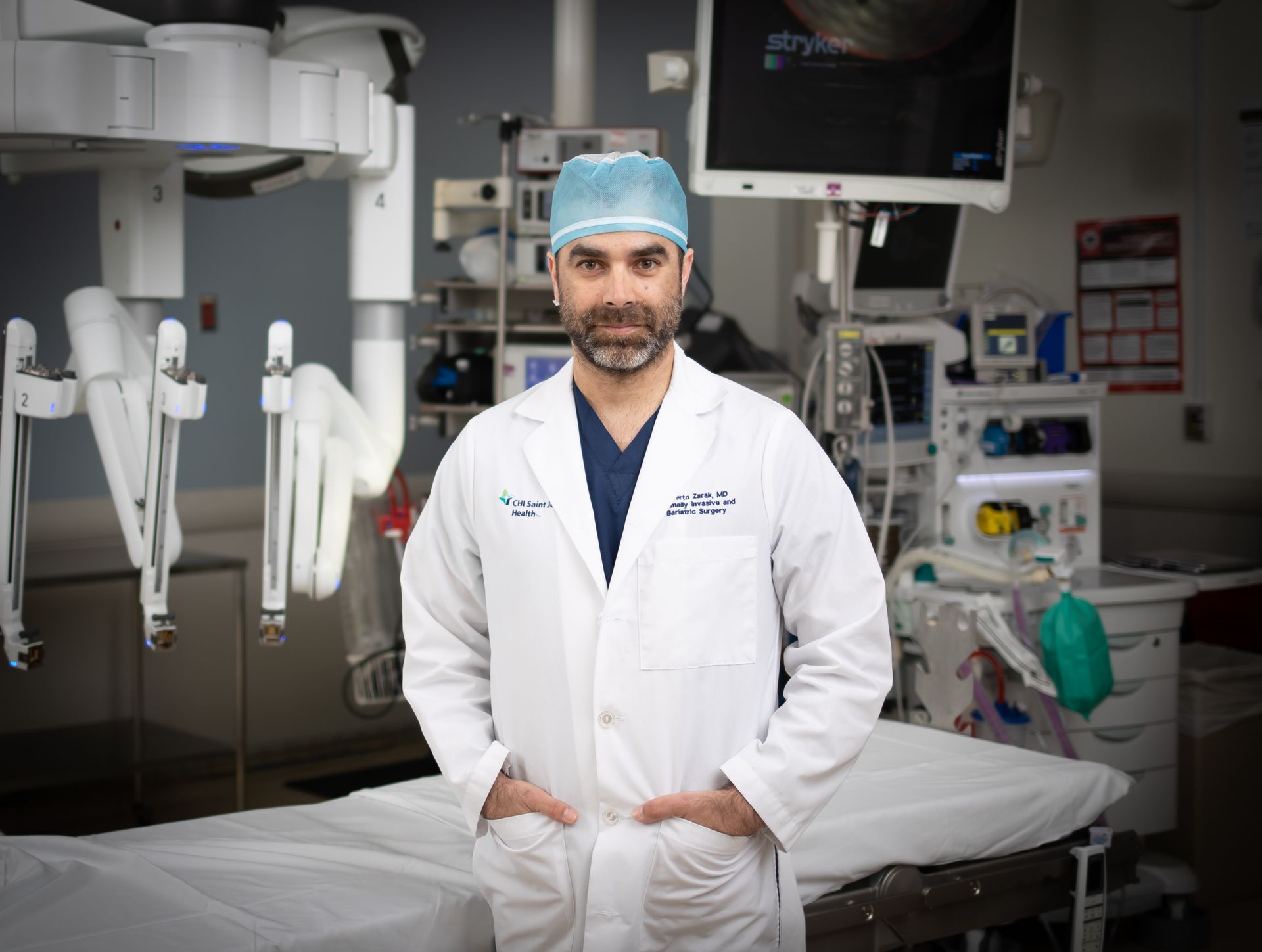 Doctor in white coat and surgical cap standing in an operating room beside medical equipment and exam table