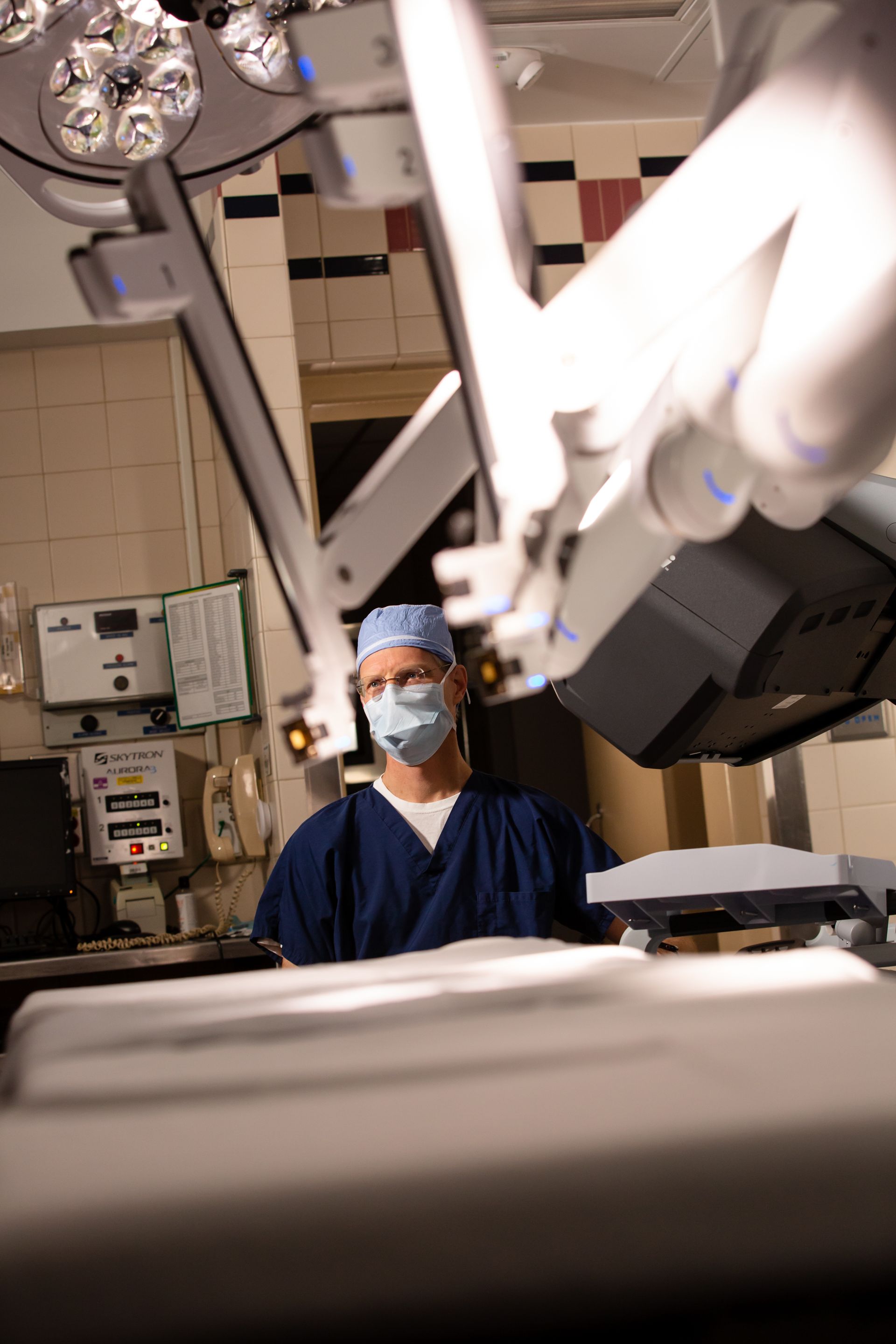 Surgeon in blue scrubs and mask beside robotic operating equipment in an operating room