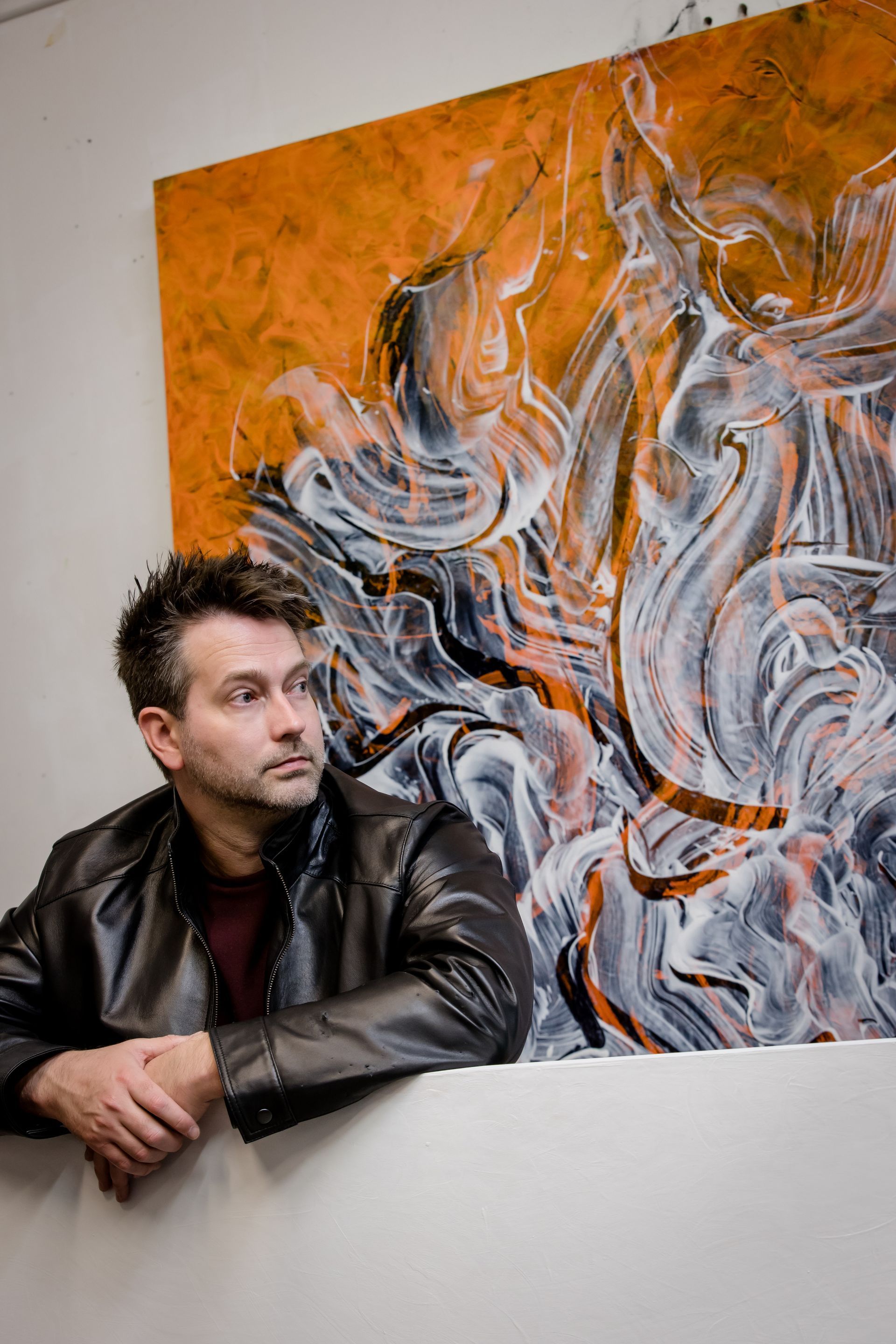 Man leaning on a white wall beside an orange abstract painting with swirling gray and black shapes