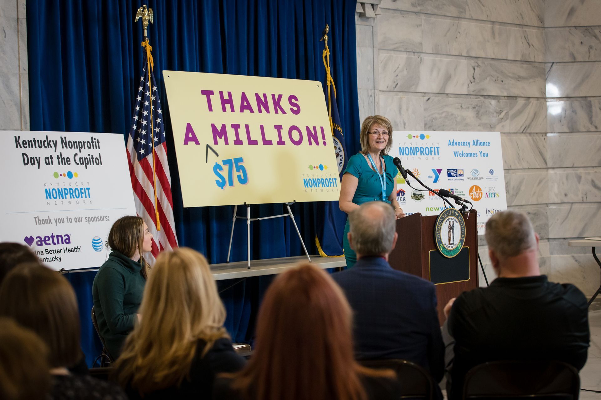Speaker at podium in teal shirt before banner reading “Thanks a million! $5” during a community event