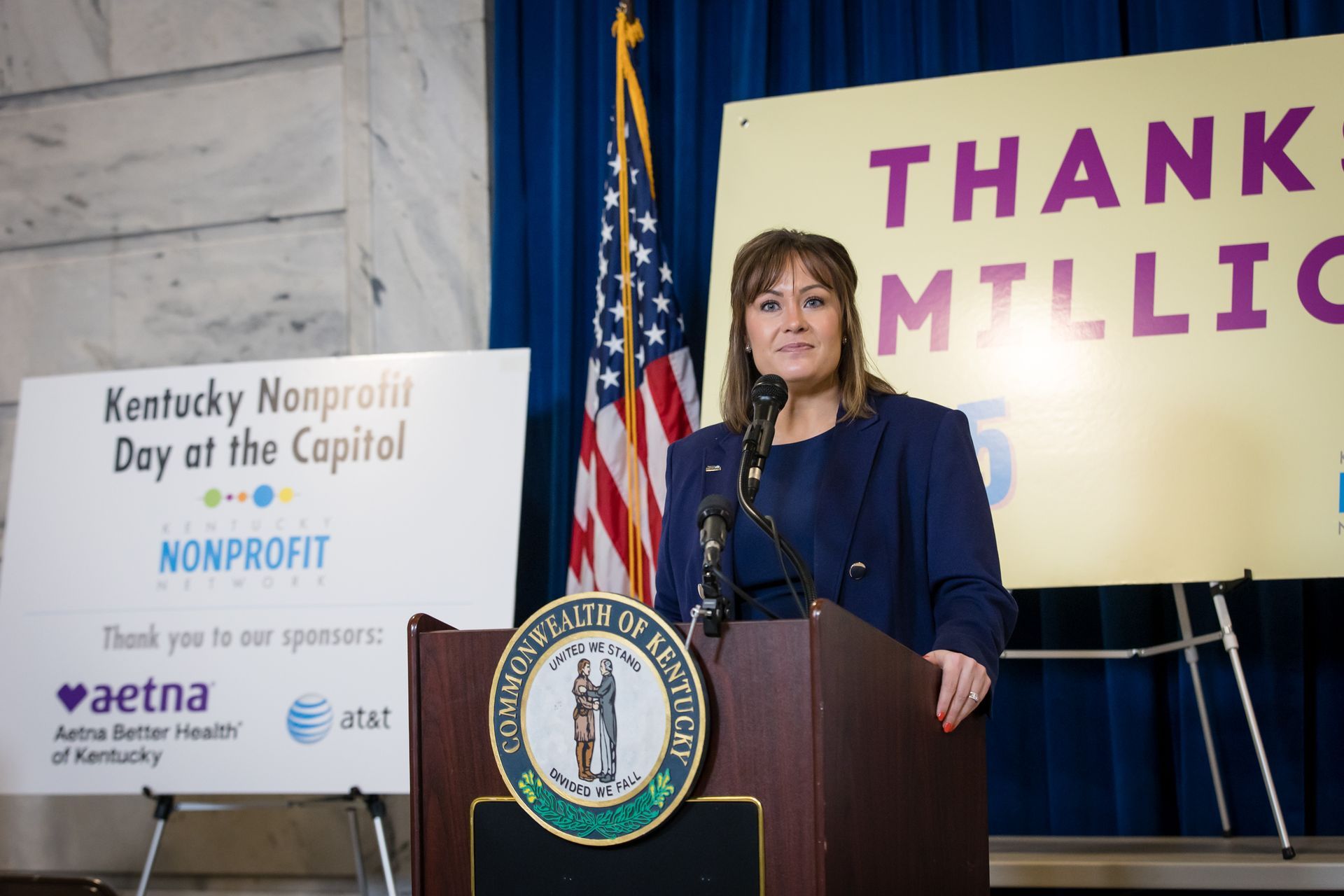Woman at podium in navy suit speaking indoors, with a “Thank you” sign and nonprofit event banner behind her