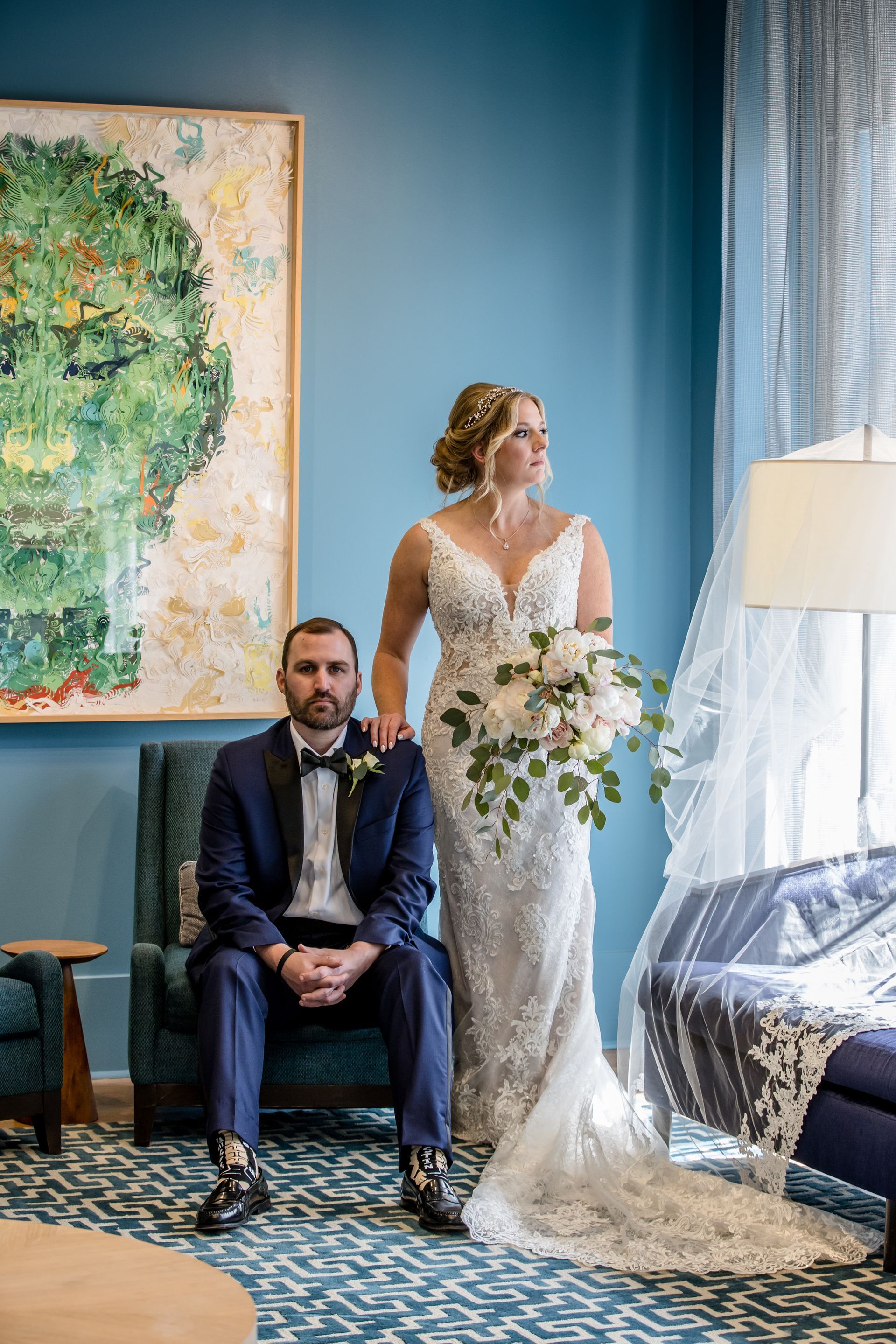 Bride in a lace gown stands by a window beside a seated groom in a blue room