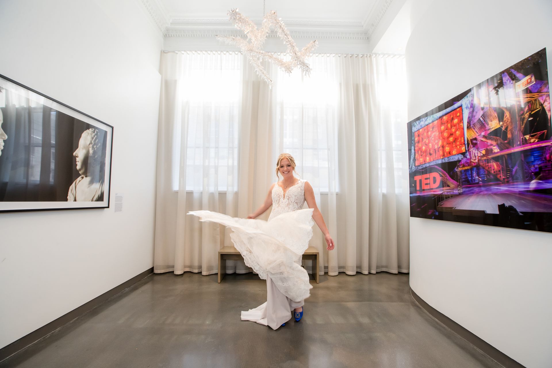 Bride in white dress posing in a bright room with curtains and wall-mounted screens.