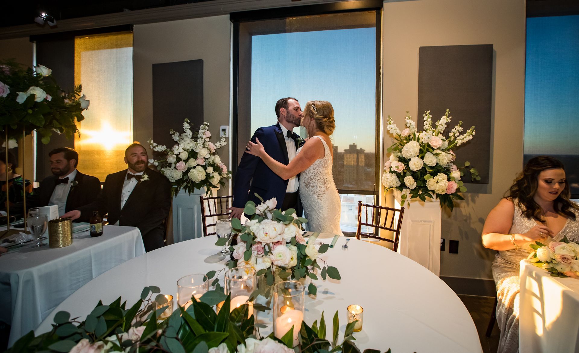 Bride and groom kissing at a wedding reception table with floral centerpieces and guests nearby