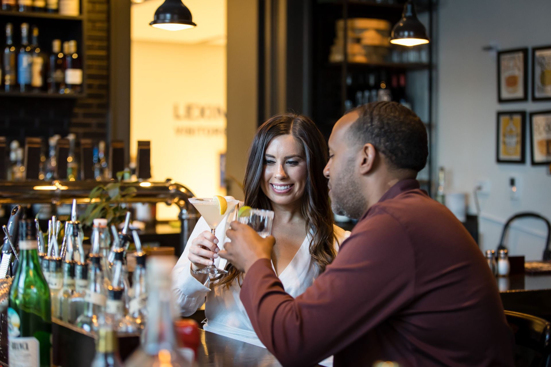 Two people toasting cocktails at a bar, smiling with bottles and hanging lights in the background
