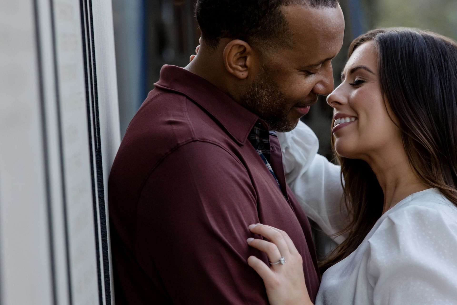 Smiling couple embracing by a window, faces close together, wearing burgundy and white tops.