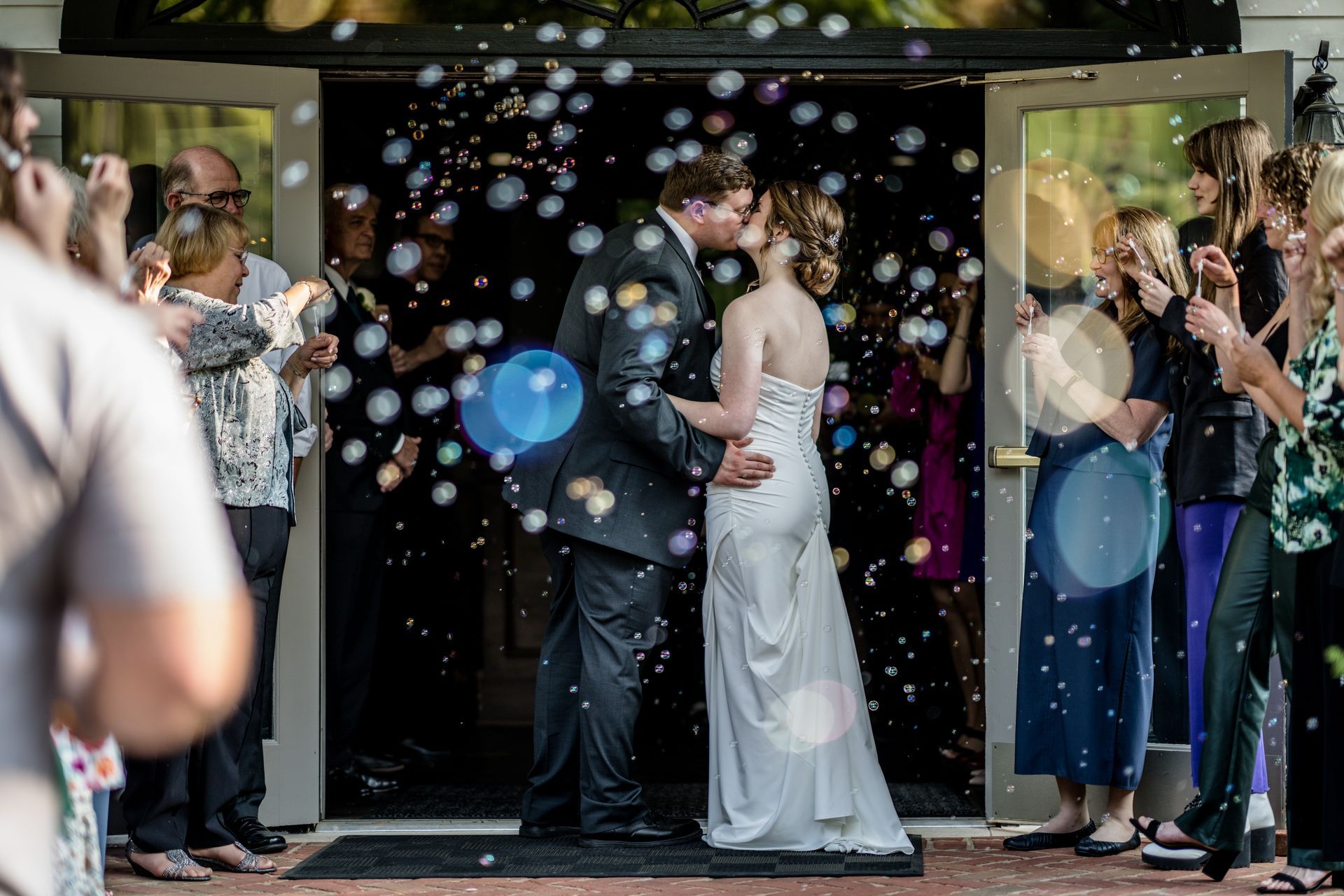 Bride and groom kissing at wedding exit as guests blow bubbles and clap