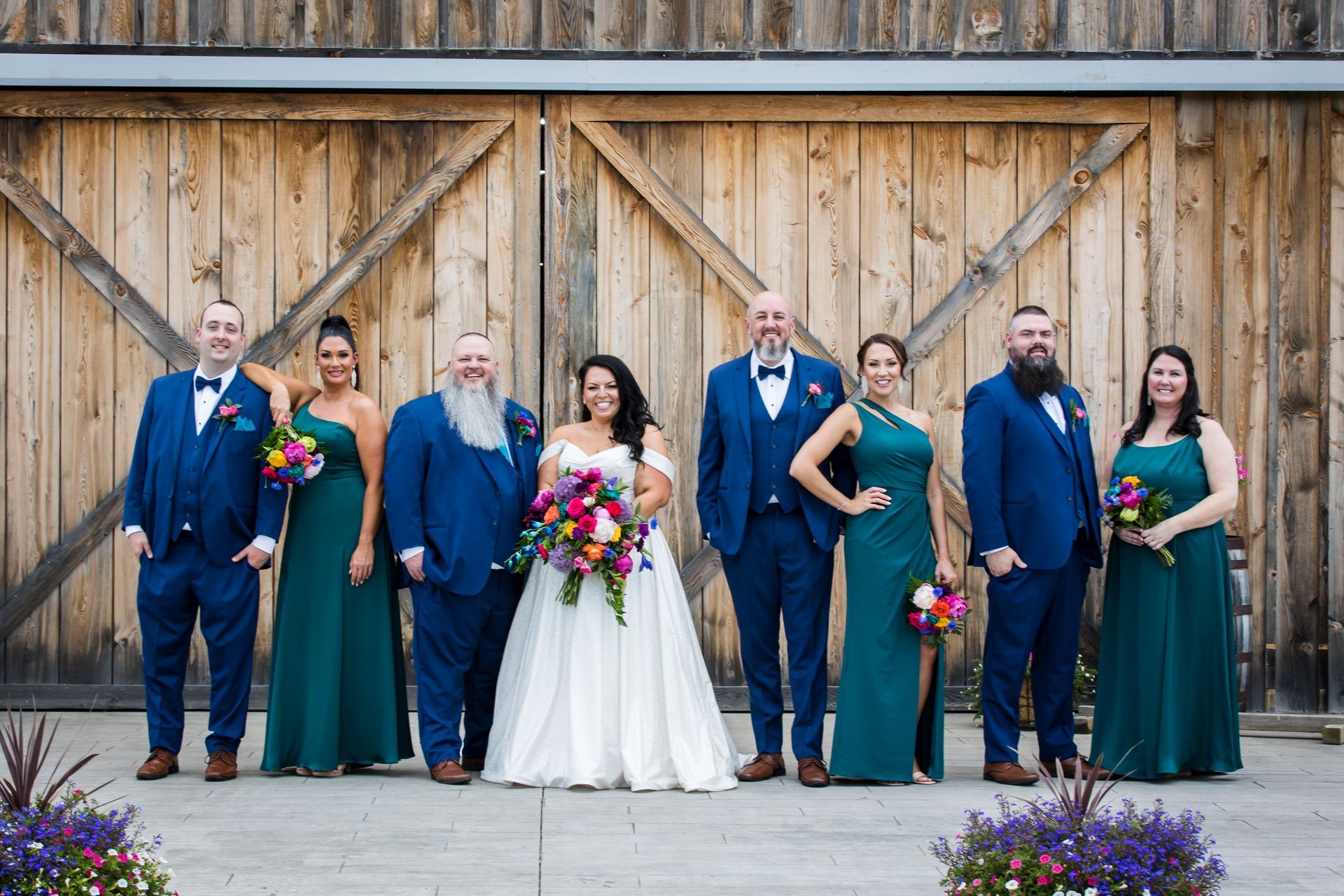 Wedding party posing in front of a wooden barn, bride in white with colorful bouquet, others in navy and green attire