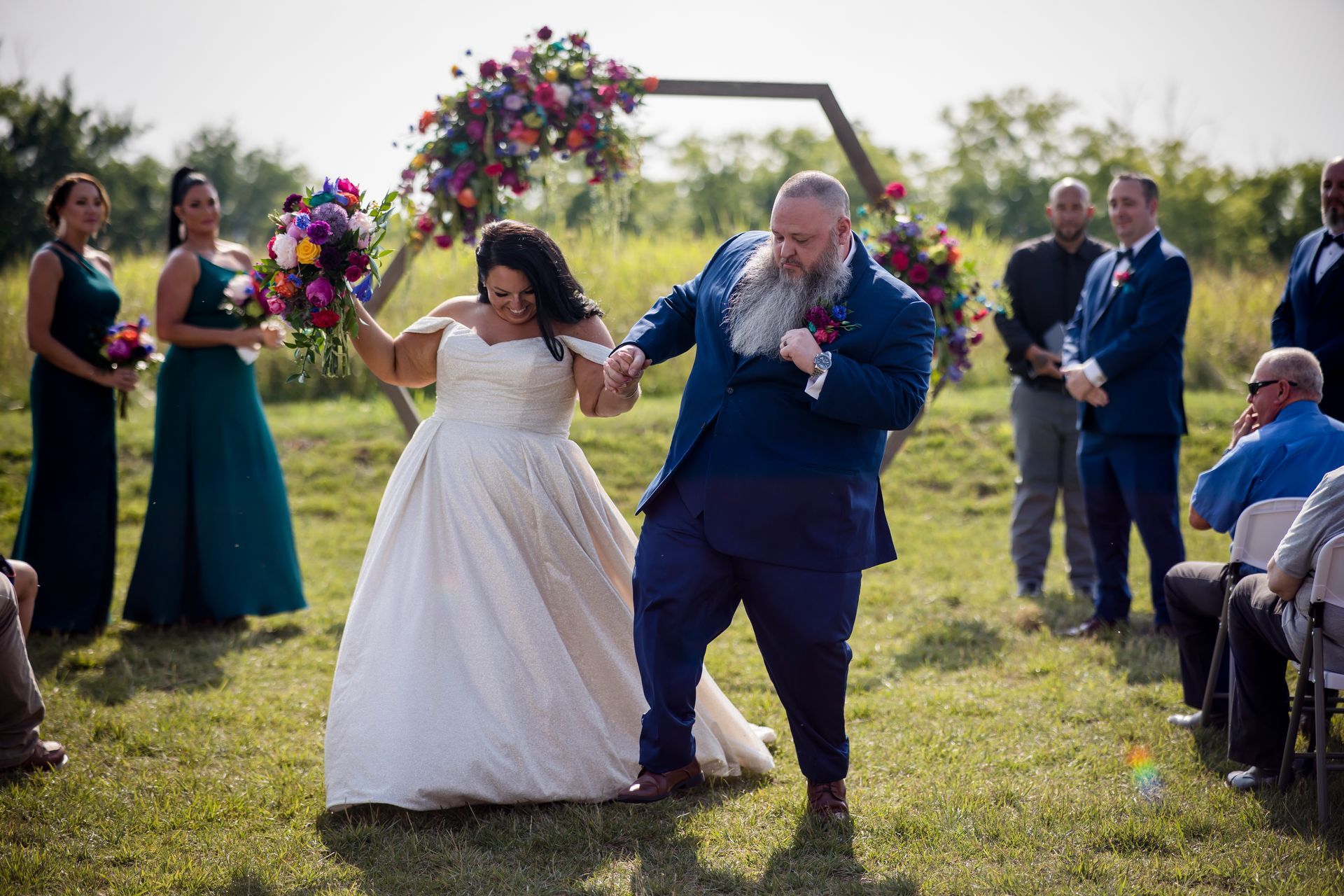 Wedding couple walking down an outdoor aisle, bride in white dress and groom in blue suit, guests nearby