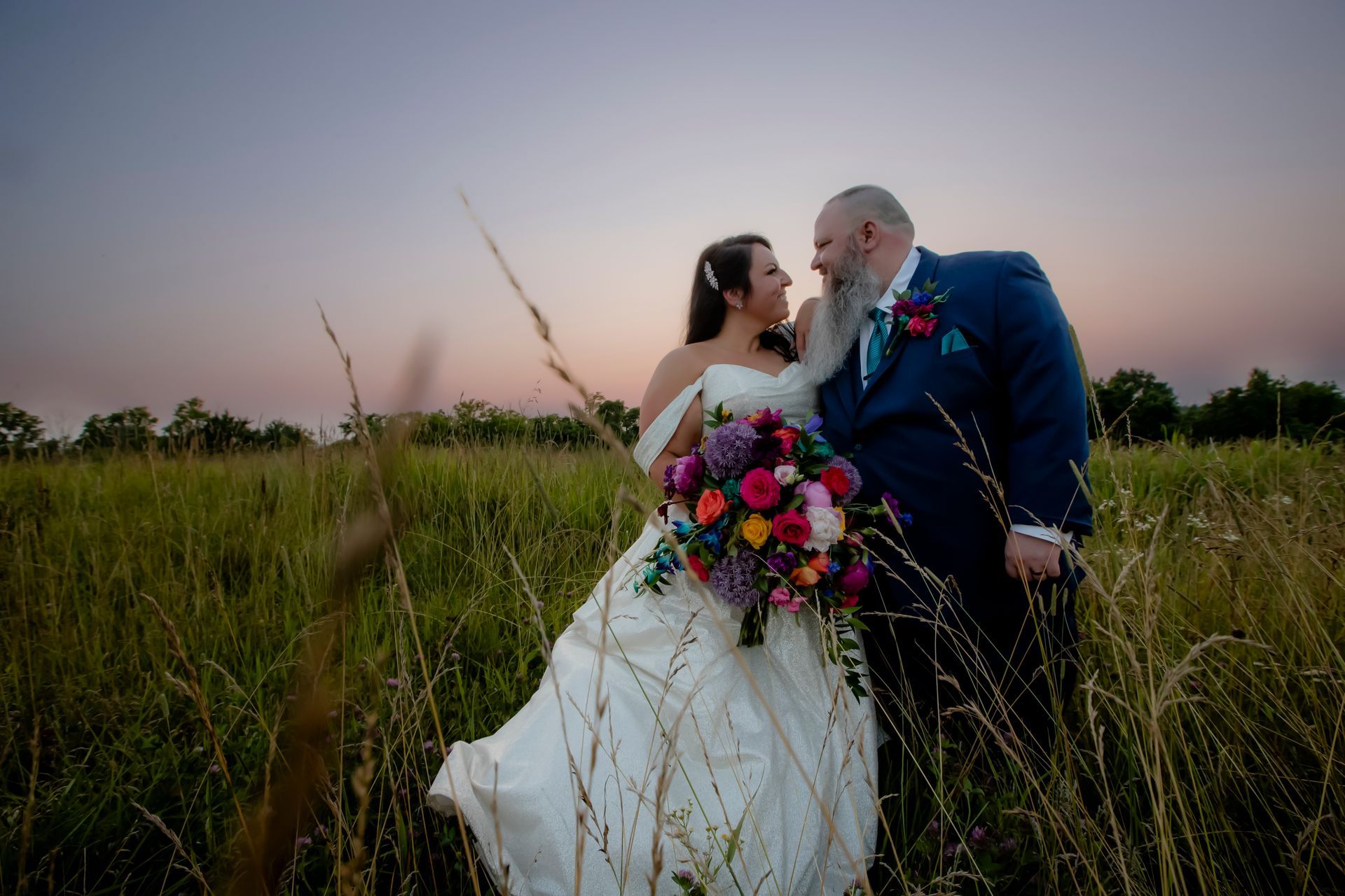 Wedding couple kissing in a grassy field at sunset, bride in white dress holding a colorful bouquet