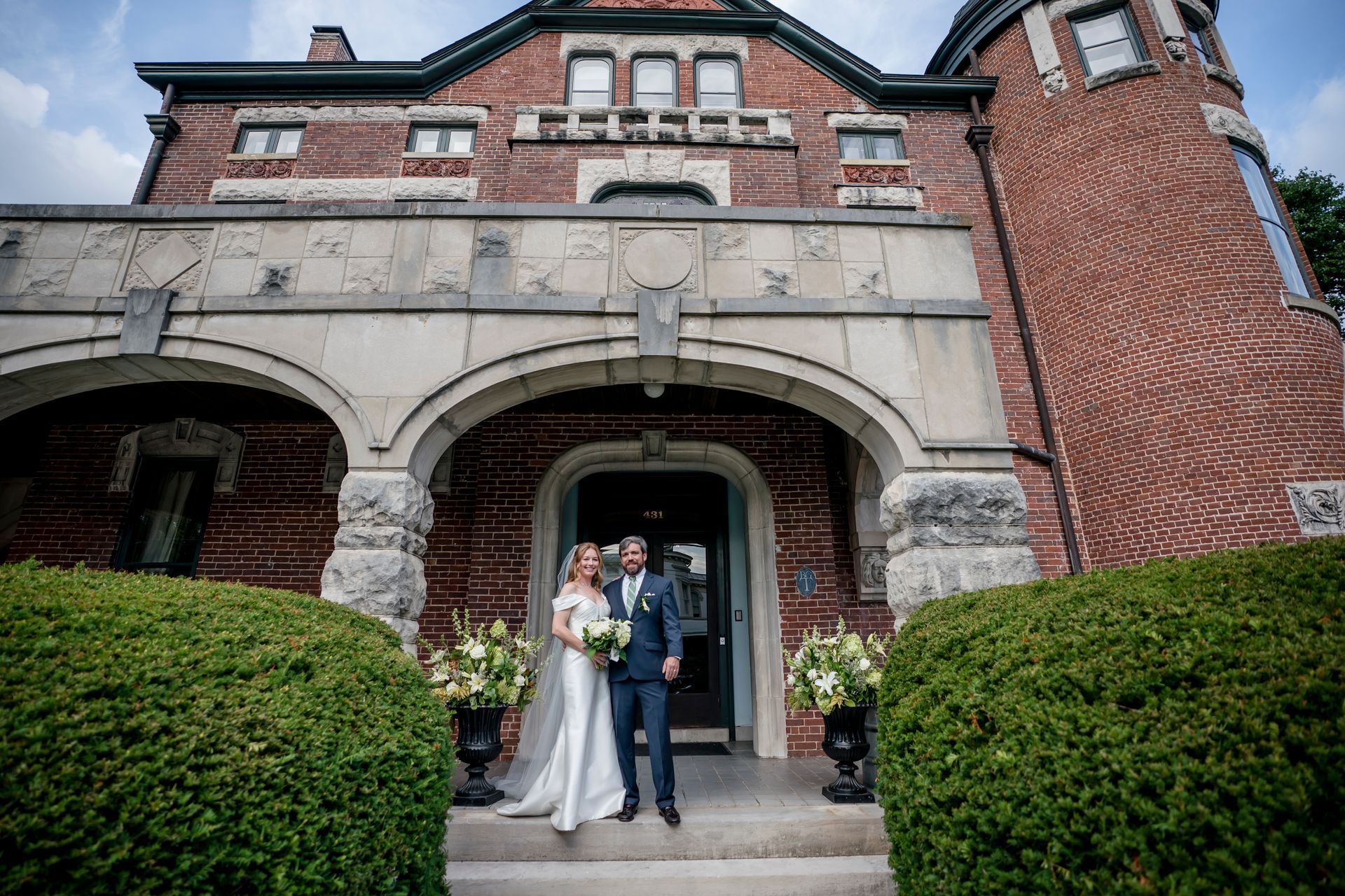 Bride and groom standing under an arched brick building entrance, holding a bouquet.