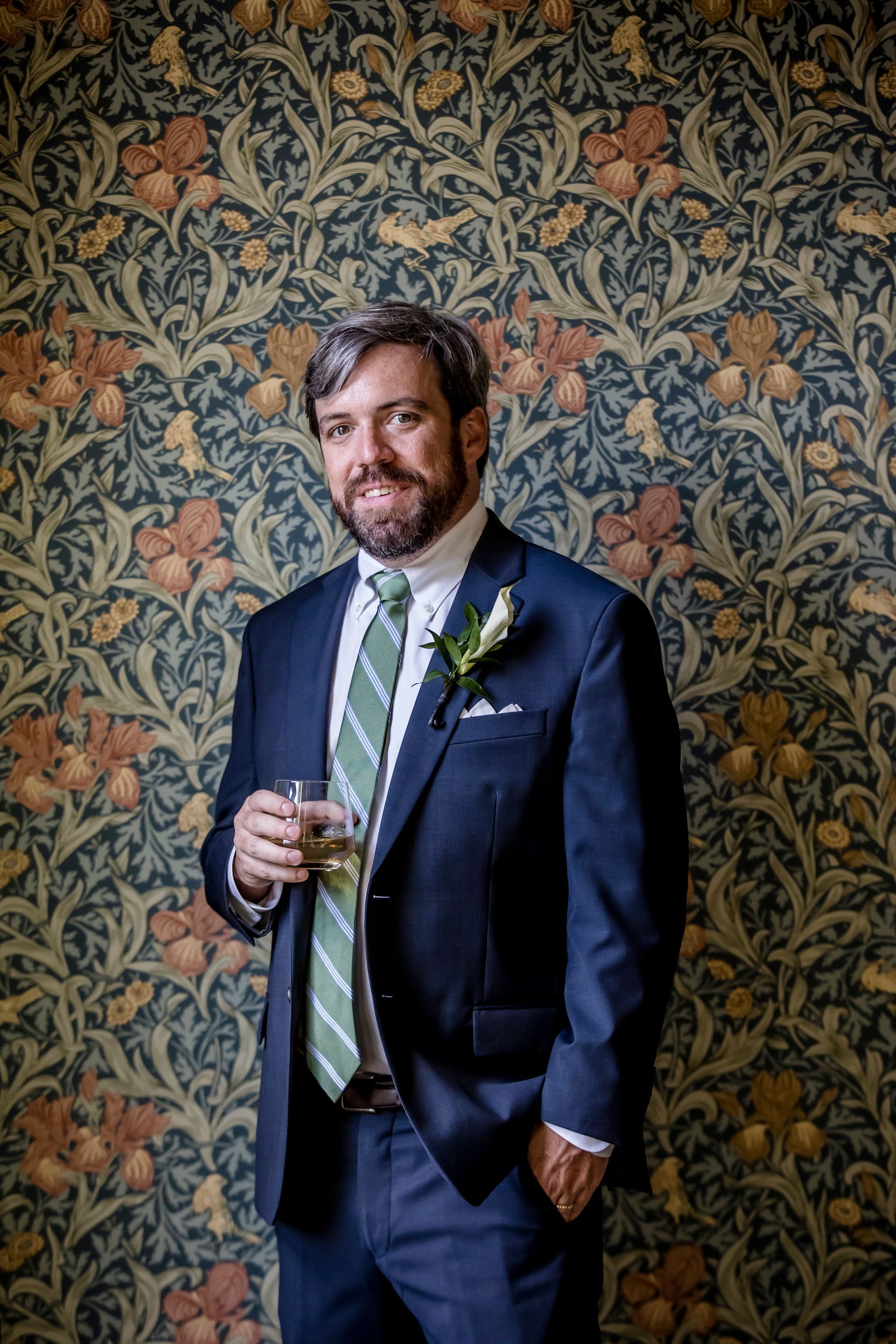 Man in navy suit holding a drink, standing before a patterned floral wallpaper backdrop.