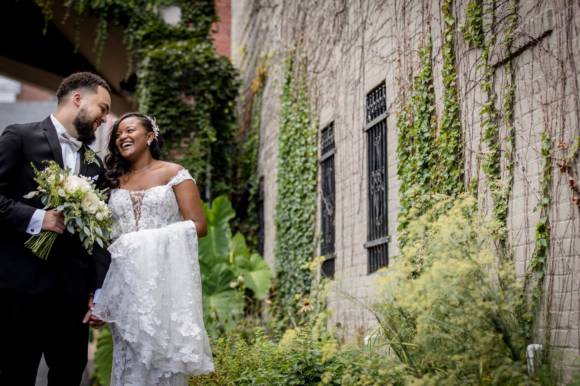 Bride and groom smiling in wedding attire beside a vine-covered stone wall
