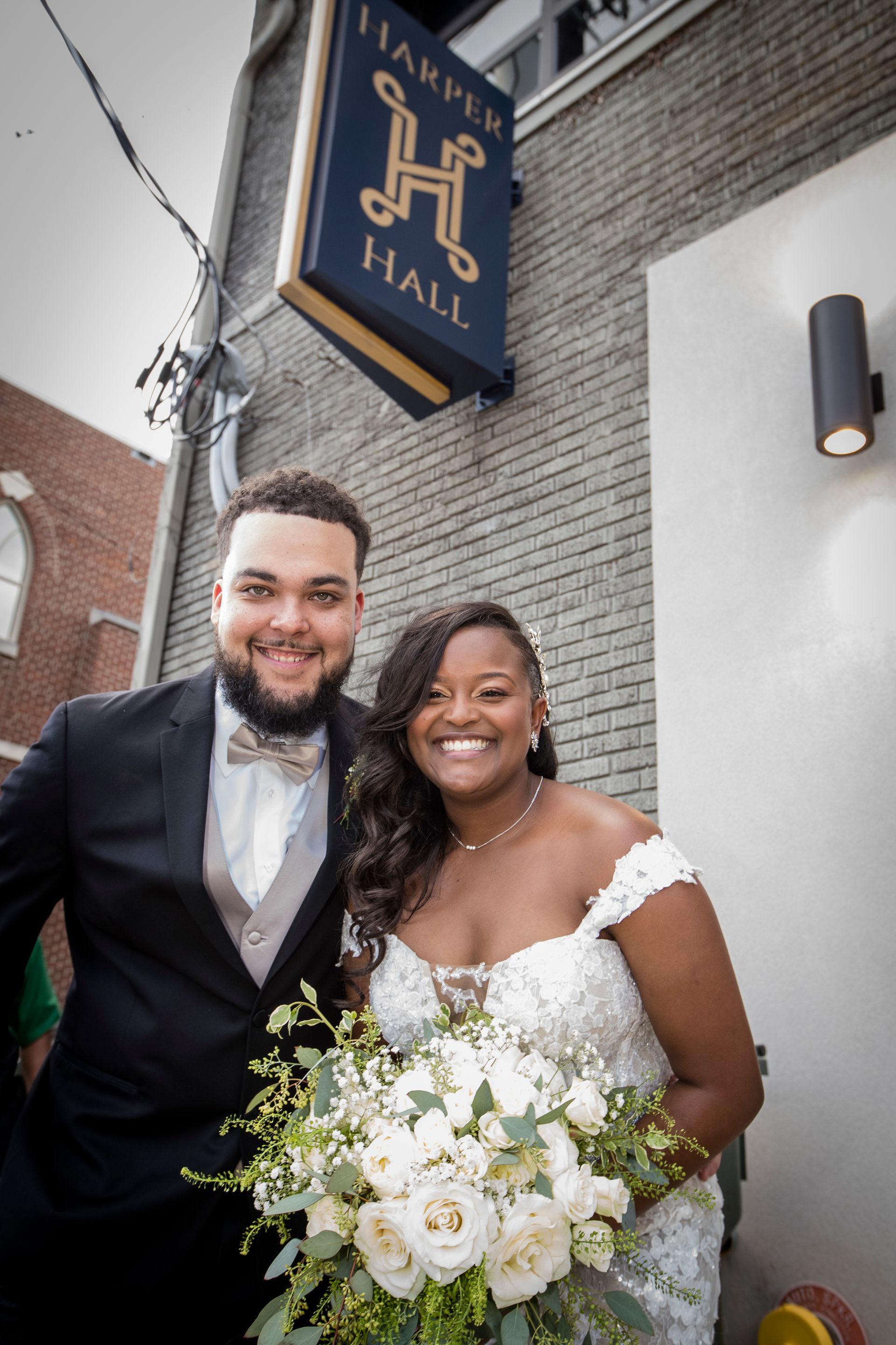 Smiling couple in formal wedding attire standing outside a hall, holding a white flower bouquet.